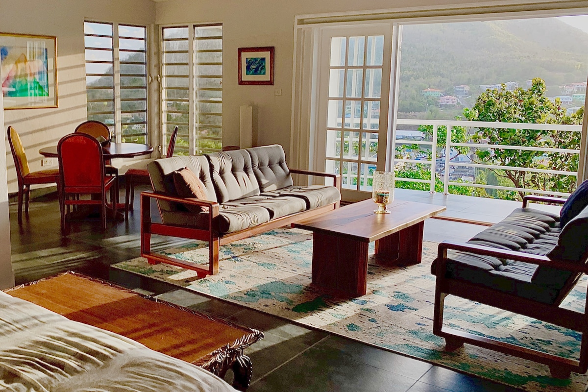 The open-plan living area features large sliding doors that welcome natural light, revealing mountain and sea views. Comfortable seating is arranged around a wooden coffee table on an area rug. A dining table with chairs is visible in the background, enhancing a spacious layout.