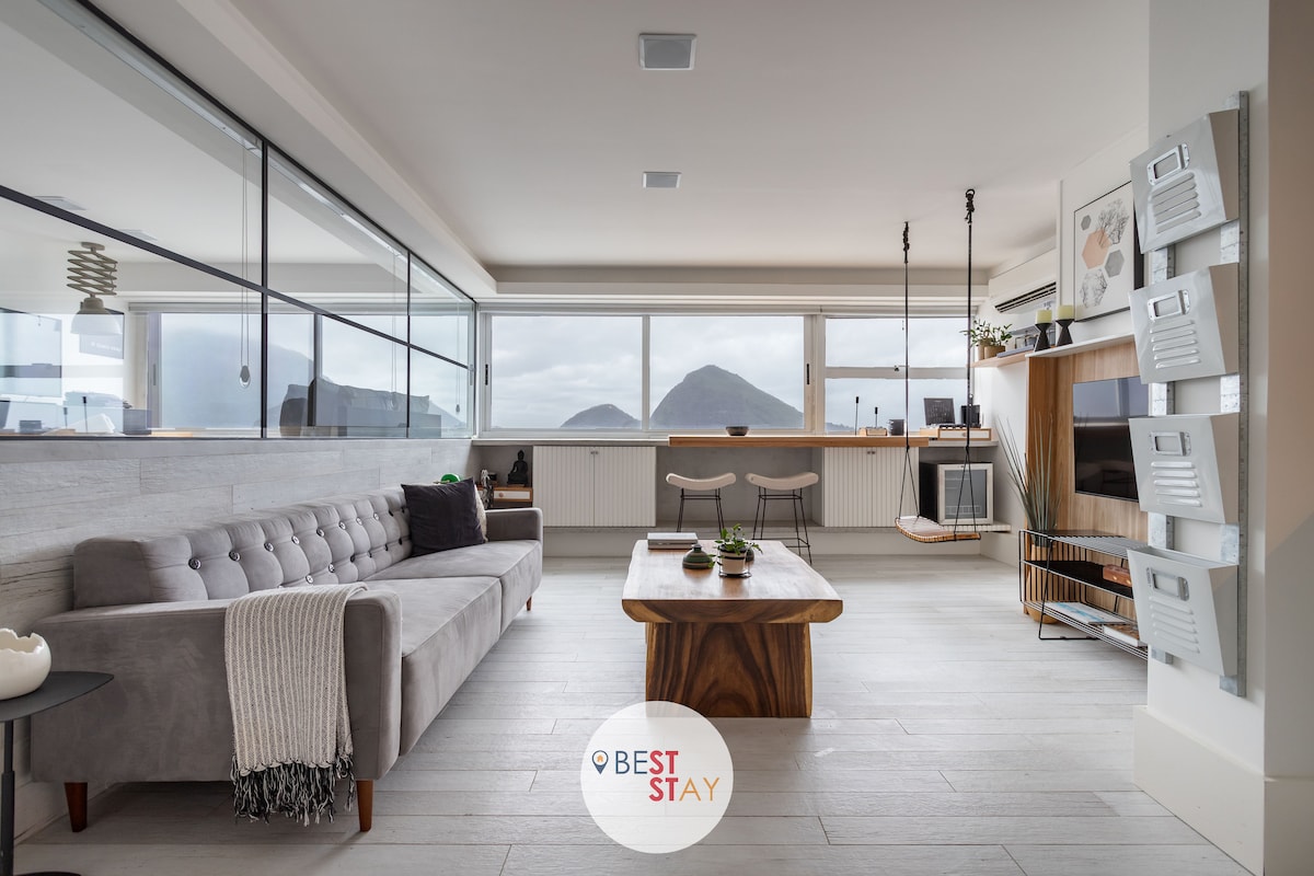 A modern living area features large windows offering views of the surrounding mountains. A spacious grey sofa is positioned alongside a wooden coffee table. A sleek television cabinet holds a Smart TV, while bar stools are placed near a counter, enhancing the room's contemporary design.