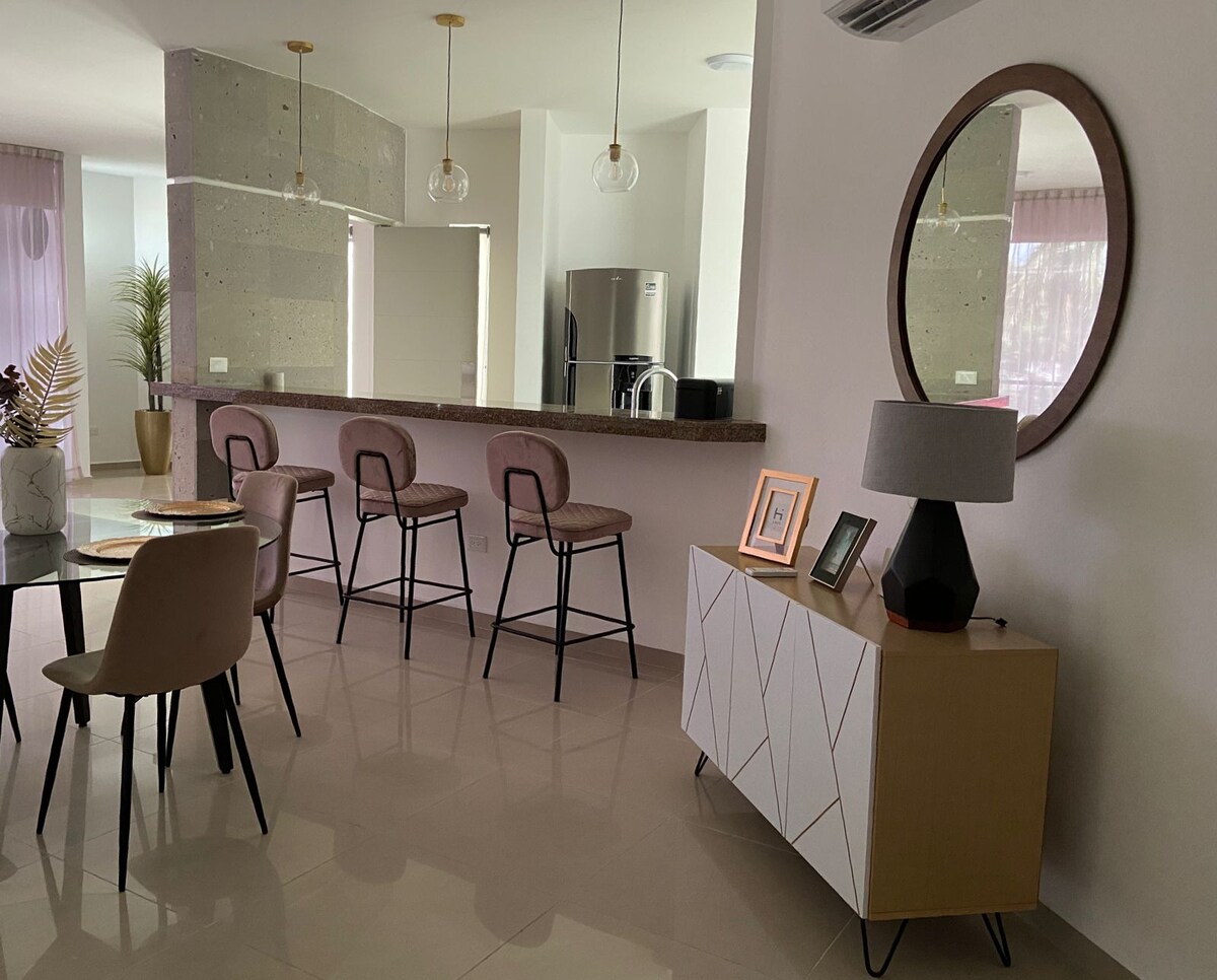 A modern dining area is visible, featuring a round table with four chairs. Behind it, a stylish bar with three stools is set against a stone wall. A decorative console table holds framed pictures and a lamp, adding to the contemporary design.