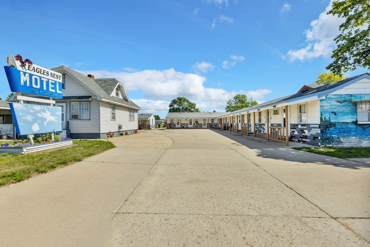 The exterior of Eagles Nest Motel features a welcoming sign and a driveway leading to the guest rooms. The building showcases a blend of vintage and modern design elements, with a white facade and decorative accents. Lush greenery and a clear blue sky enhance the serene atmosphere.
