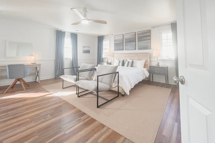 Welcome to the expansive primary bedroom, featuring a luxurious king bed, cozy seating area, smart TV, desk, and an en suite bath with a walk-in closet, all enveloped in calming light blue and white hues and crisp white linens for a serene escape.