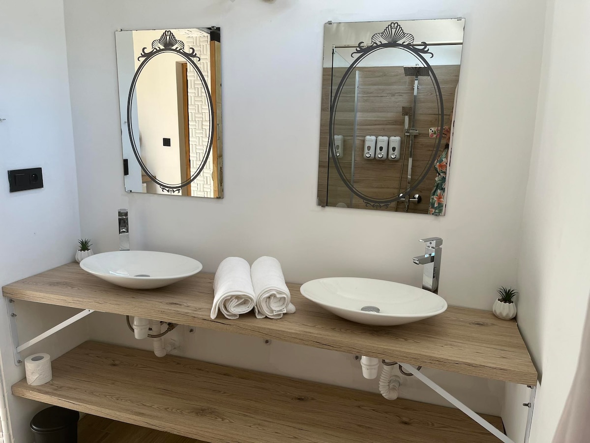 A modern bathroom features two elegant sinks situated on a wooden countertop. Above each sink, round mirrors are framed in decorative metal. Fresh towels are neatly rolled and placed between the sinks, accompanied by small decorative plants on each side.