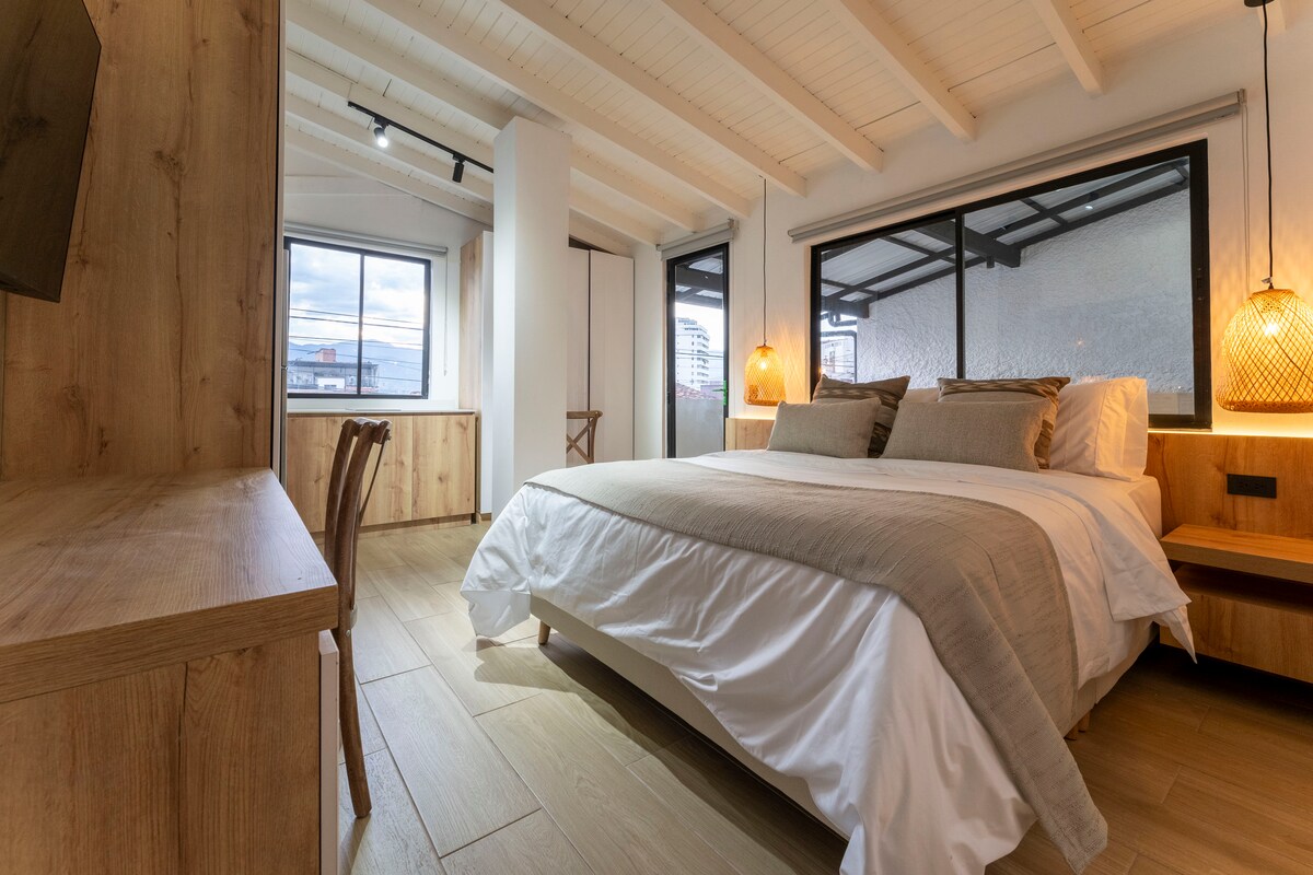 A spacious sleeping area features a large bed with a soft duvet, complemented by two decorative pillows. Natural light enters through the expansive windows, illuminating the wooden furnishings, including a desk and a chair. Subtle light fixtures add a touch of warmth to the room.