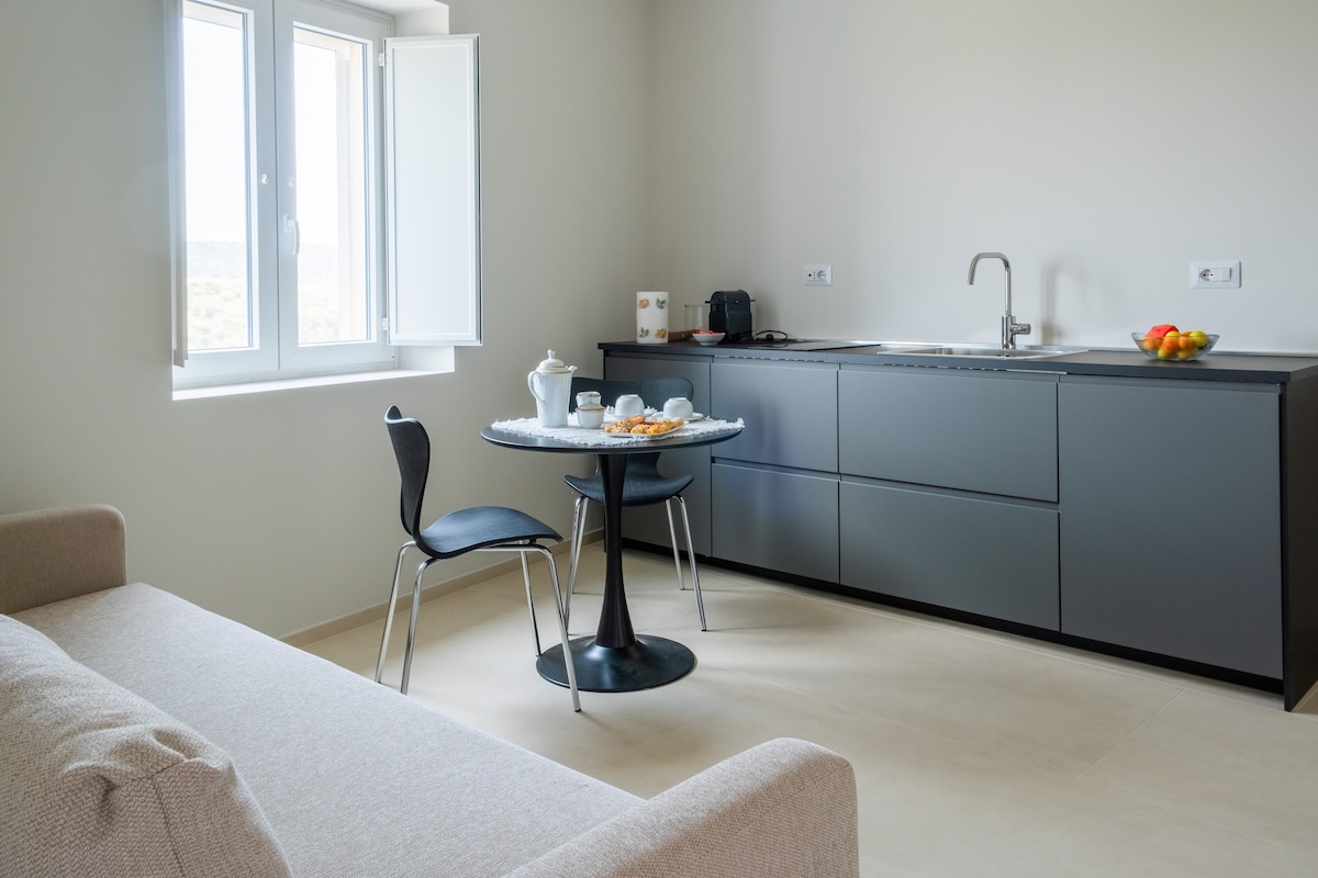 A contemporary kitchen area is seen, featuring sleek cabinetry and a round dining table with two chairs. Sunlight enters through a pair of open windows, illuminating a simple setup of tea and snacks on the table, while a cozy sofa is positioned nearby.