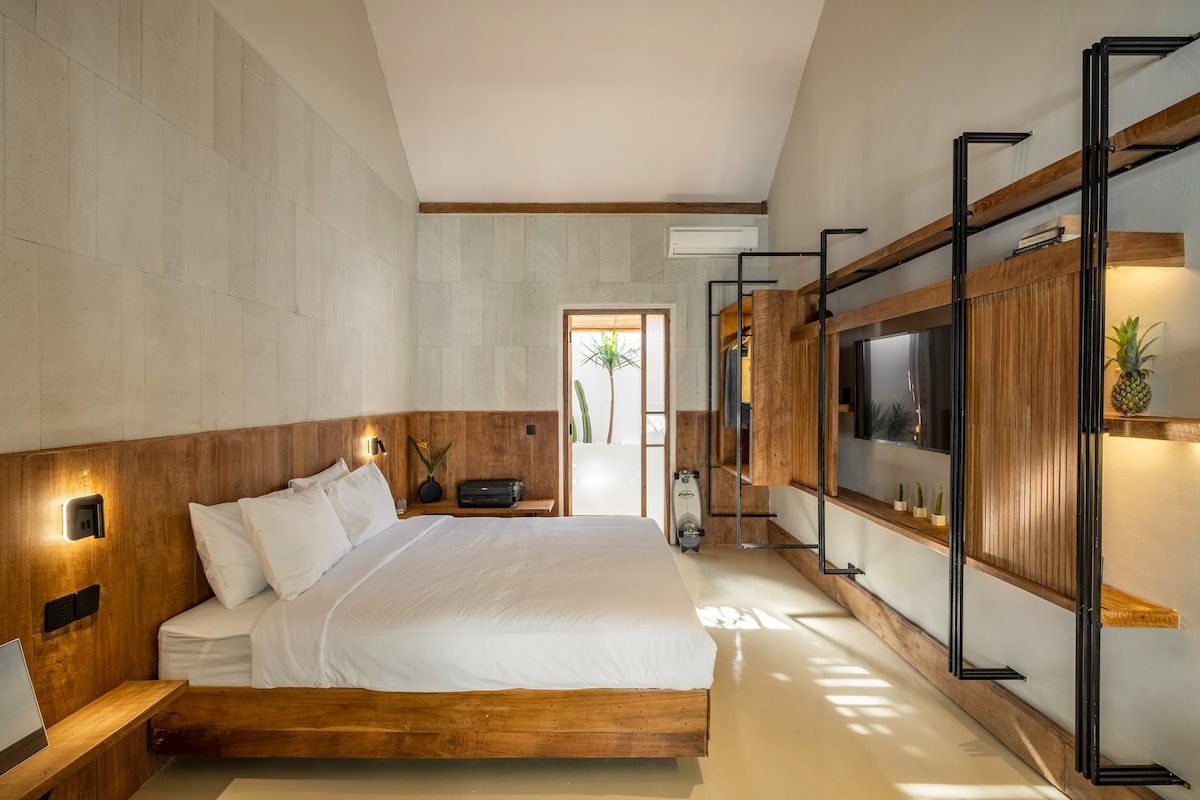 This spacious suite features a cozy king-size bed positioned against a wooden accent wall. Natural light filters through the windows, highlighting minimalist shelving and a modern television setup. The serene atmosphere is enhanced by neutral tones and tropical decor, inviting relaxation.