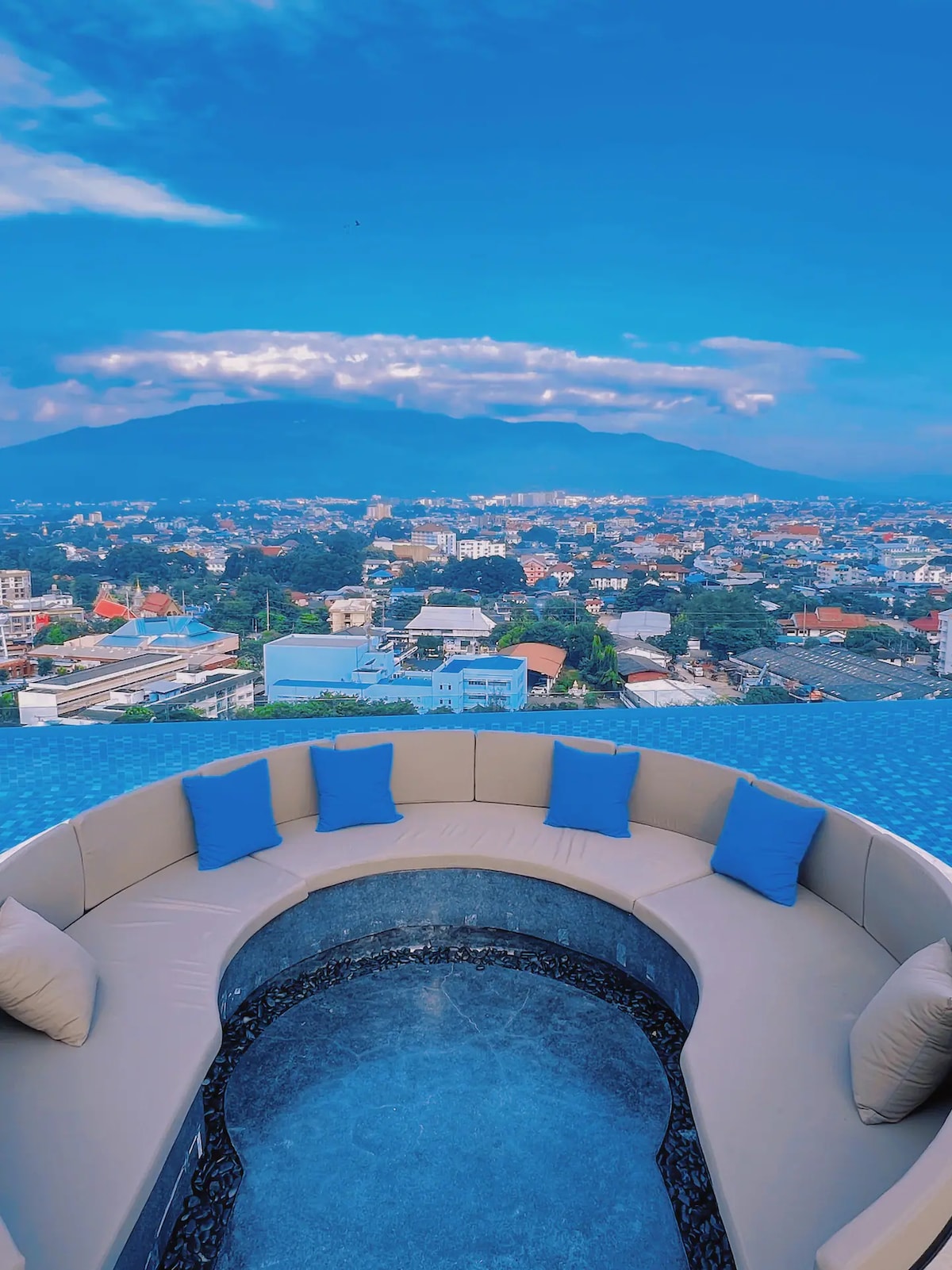 A round seating area is positioned next to the infinity pool, overlooking a panoramic view of the city and mountains. Soft cushions in vibrant blue provide comfort, while the sky reflects shades of blue and white, enhancing the serene atmosphere.