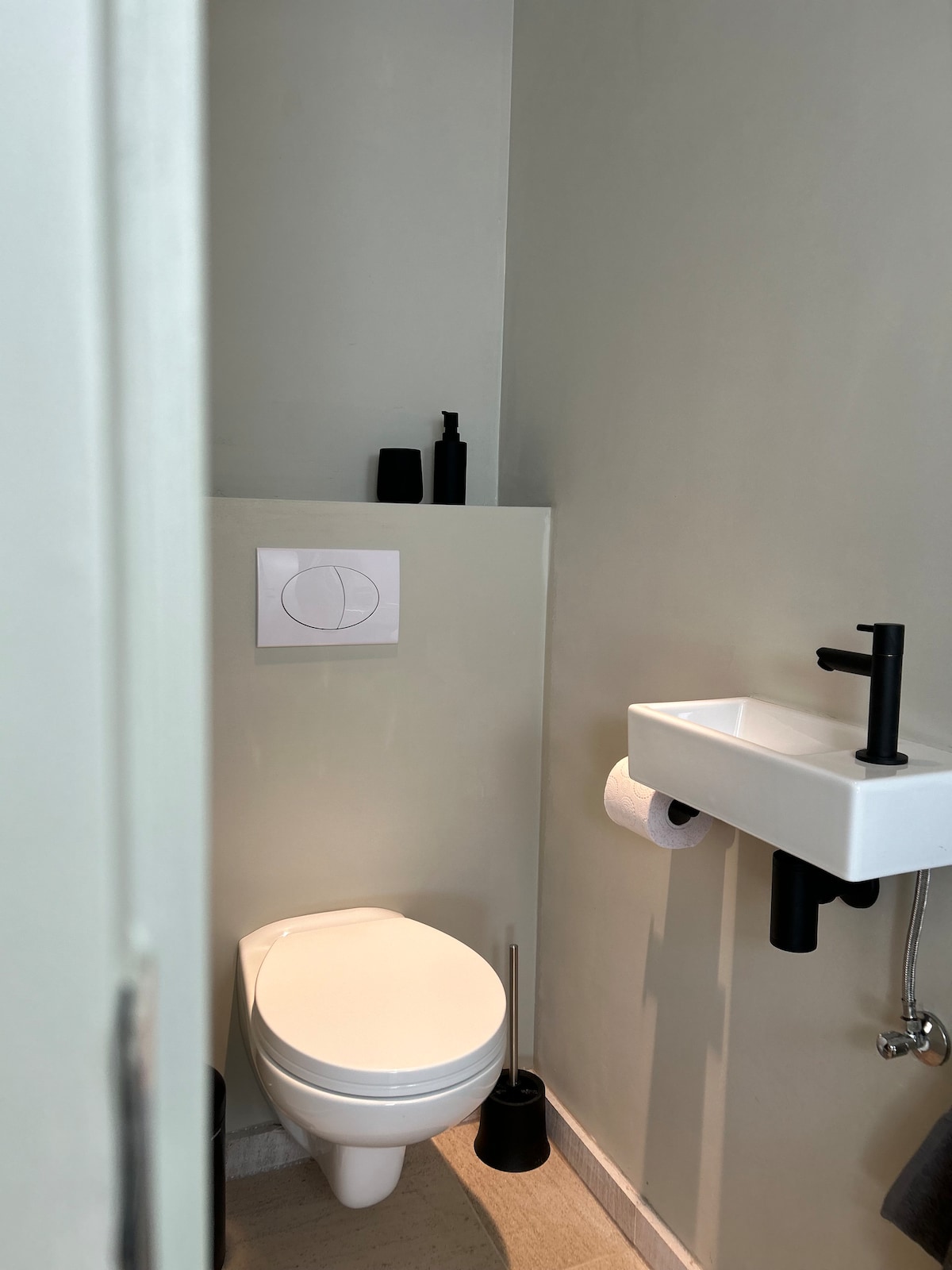 The bathroom features a modern toilet and a compact sink mounted on the wall. Essentials such as a toilet paper holder and a soap dispenser are present, enhancing convenience. Neutral-colored walls contribute to a clean and minimalistic aesthetic.