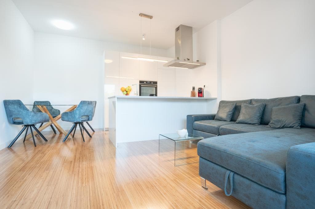 A modern living space is presented, featuring a comfortable sectional sofa in soft grey. A small glass table complements the dining area, which includes two blue upholstered chairs. The kitchen area, equipped with sleek appliances and a minimalist design, is visible in the background.