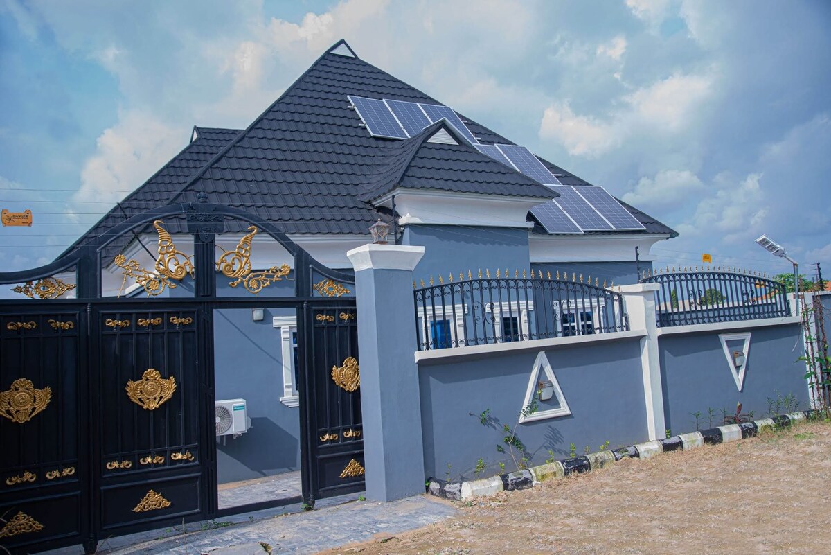 The exterior of a modern house is showcased, featuring a dark roof and light blue walls. Solar panels are installed on the roof, and an ornate black gate and fence enhance the property's entrance. A well-maintained yard is visible, complementing the overall aesthetic.