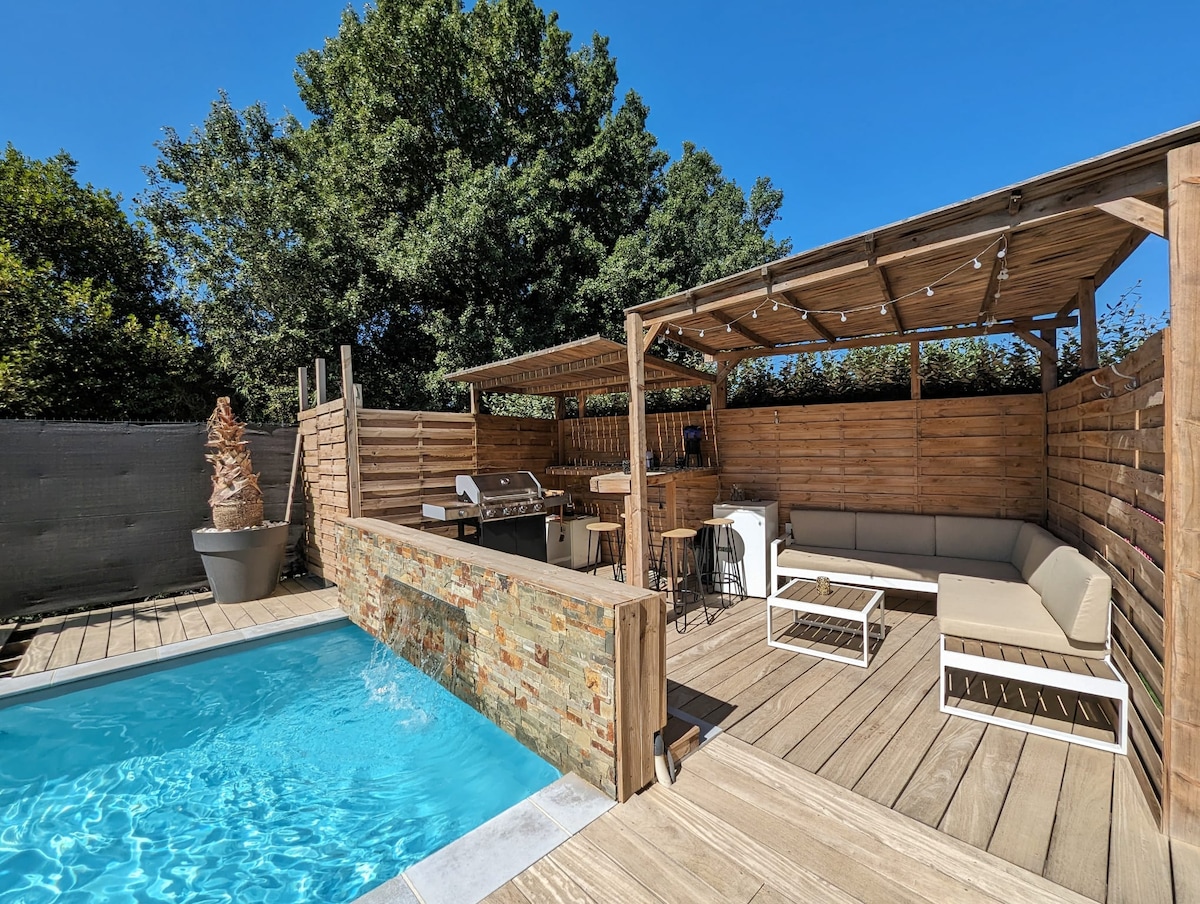 An outdoor area features a private pool alongside a wooden deck. A covered lounge space with comfortable seating is paired with a grill setup, providing an ideal space for al fresco dining. Lush greenery surrounds the area, creating a serene environment.