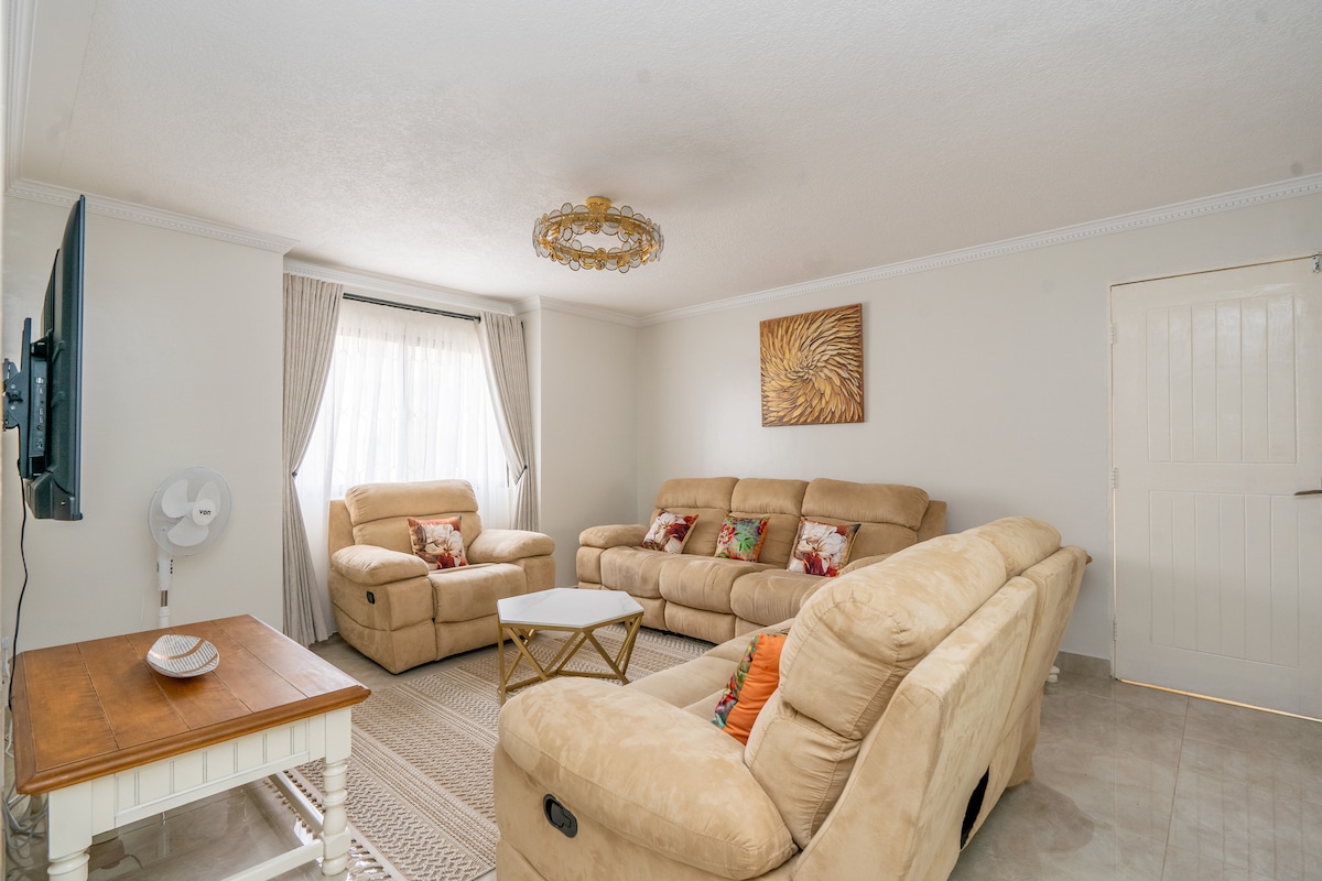 A comfortable living room is presented with soft beige sofas adorned with decorative cushions. A small coffee table sits in the center, and large windows offer natural light, complemented by sheer curtains. A unique golden wall art adds a touch of elegance to the space.