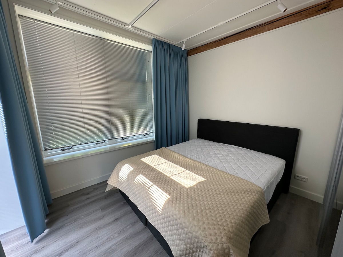 A bright sleeping area is featured, with a spacious bed dressed in a light quilt. Natural light filters through a partially opened window, complemented by soft blue curtains. Light wood accents on the walls add warmth to the contemporary design.