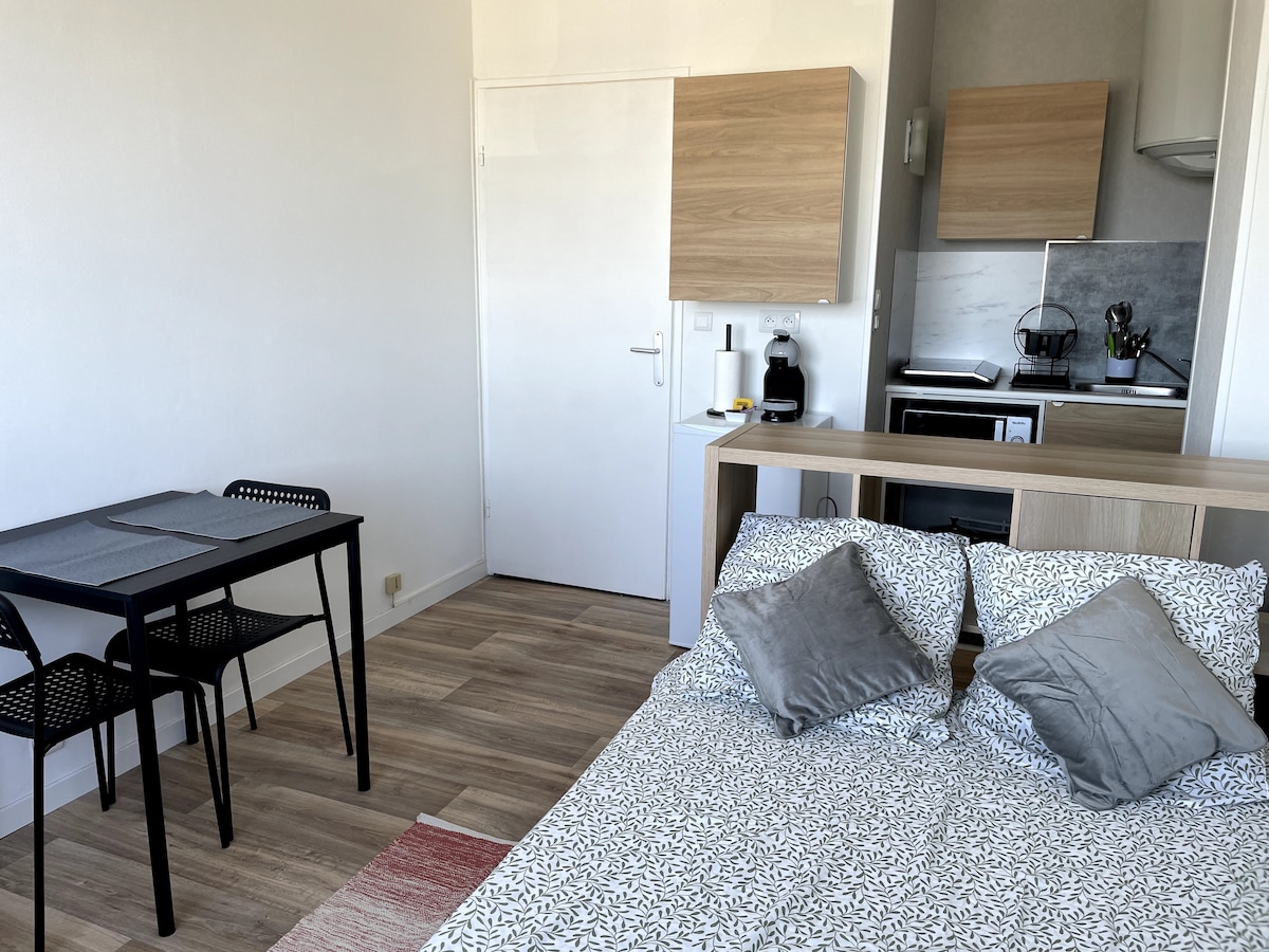 A bright studio space features a spacious bed adorned with patterned bedding and decorative pillows. A minimalist dining area is equipped with a small table and two chairs. The kitchenette includes modern appliances, offering a functional and inviting atmosphere.