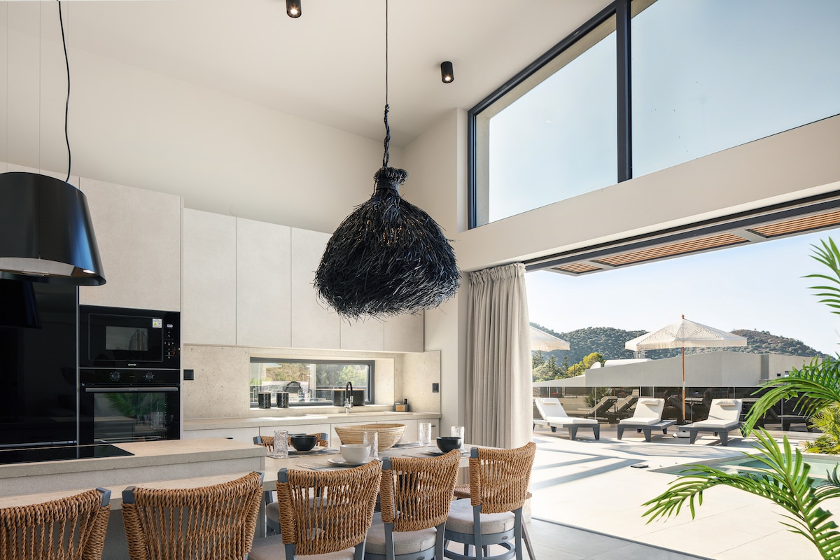 A spacious open-plan kitchen and dining area features modern appliances and a large dining table. Stylish wicker chairs surround the table, complemented by a statement pendant light. Floor-to-ceiling glass doors lead to an outdoor terrace, enhancing the connection with the landscape.