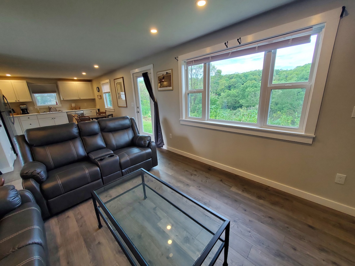 A living room is depicted with ample natural light streaming through a large picture window that frames views of the wooded area outside. A glass coffee table is placed in front of a reclining sofa and love seat, both featuring a dark upholstery.