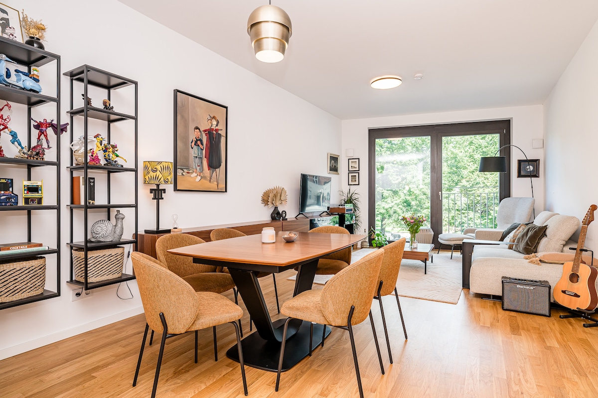 A dining area showcases a wooden table surrounded by four upholstered chairs. A bookshelf filled with decorative items and toys lines one wall, while a comfortable seating area with a light-colored couch and an acoustic guitar is visible. Large windows offer views of greenery.