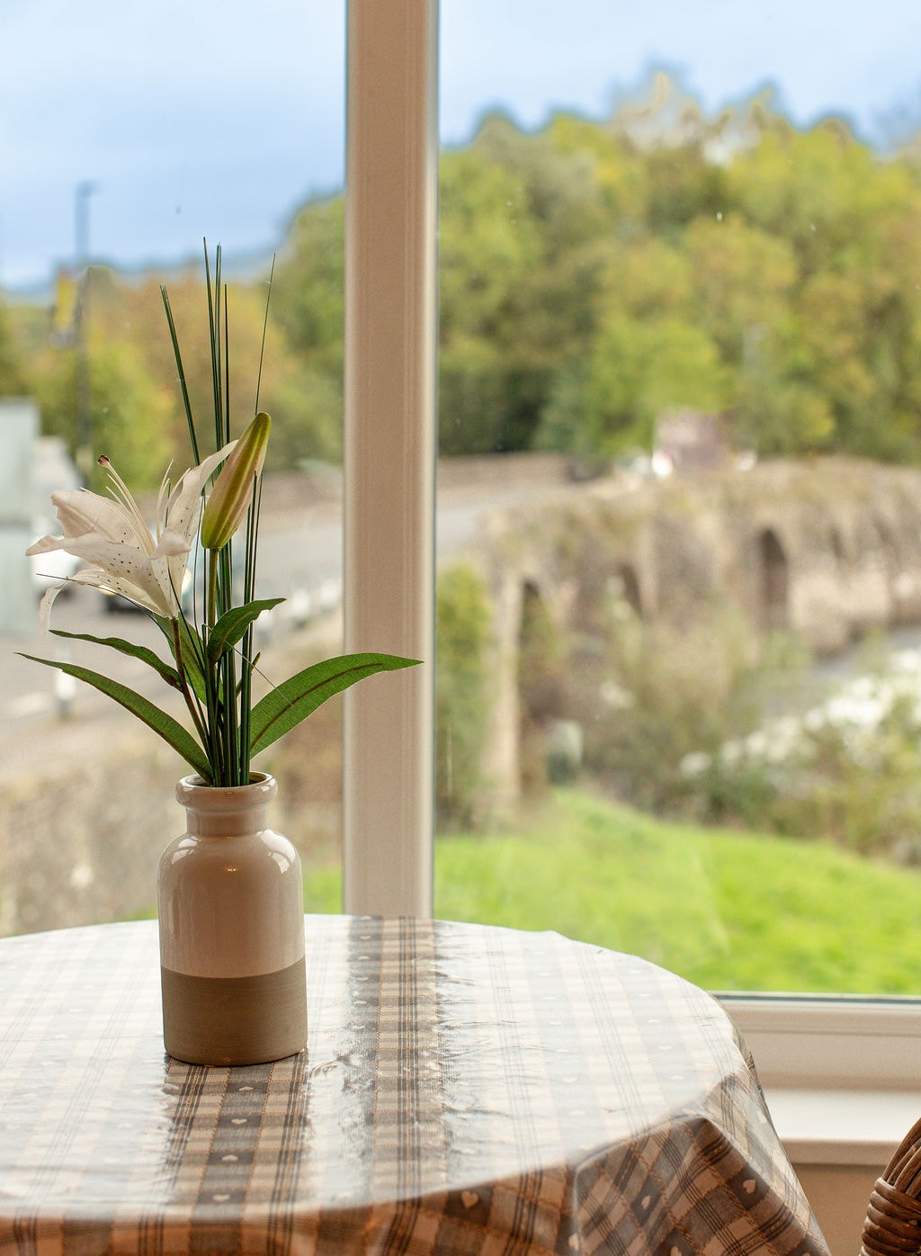 A small table is situated near a window, displaying a vase with fresh lilies. The background features greenery and a stone bridge, visible through the window. The table is covered with a checkered cloth, enhancing the tranquil setting.