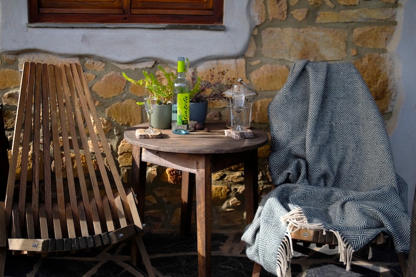 A cozy outdoor seating area is depicted, featuring a round wooden table surrounded by two chairs. One chair displays a soft blanket draped over it, while a decorative vase with greenery and a bottle are placed on the table. A stone wall serves as a rustic backdrop.