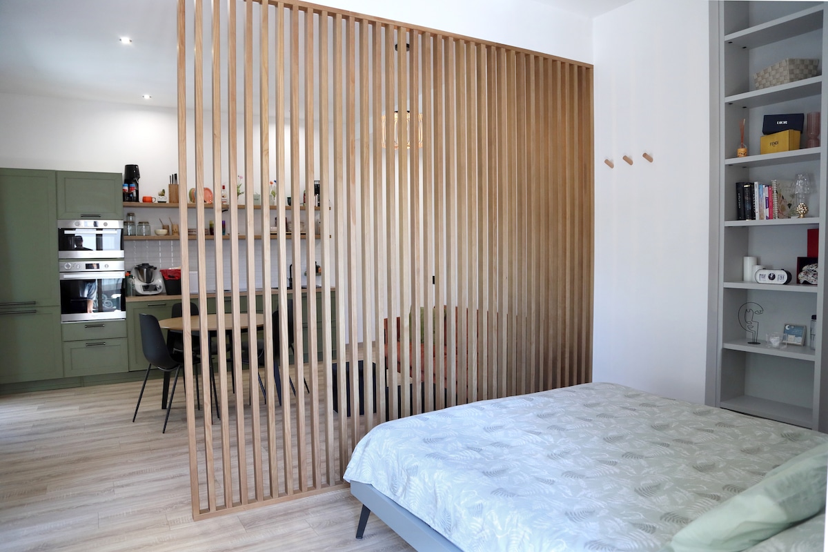 A wooden slat partition defines the sleeping area, separating it from the kitchen and dining space. A spacious bed is positioned in front, while the open kitchen area and dining chairs are visible in the background, providing a sense of organization within the monolocale.