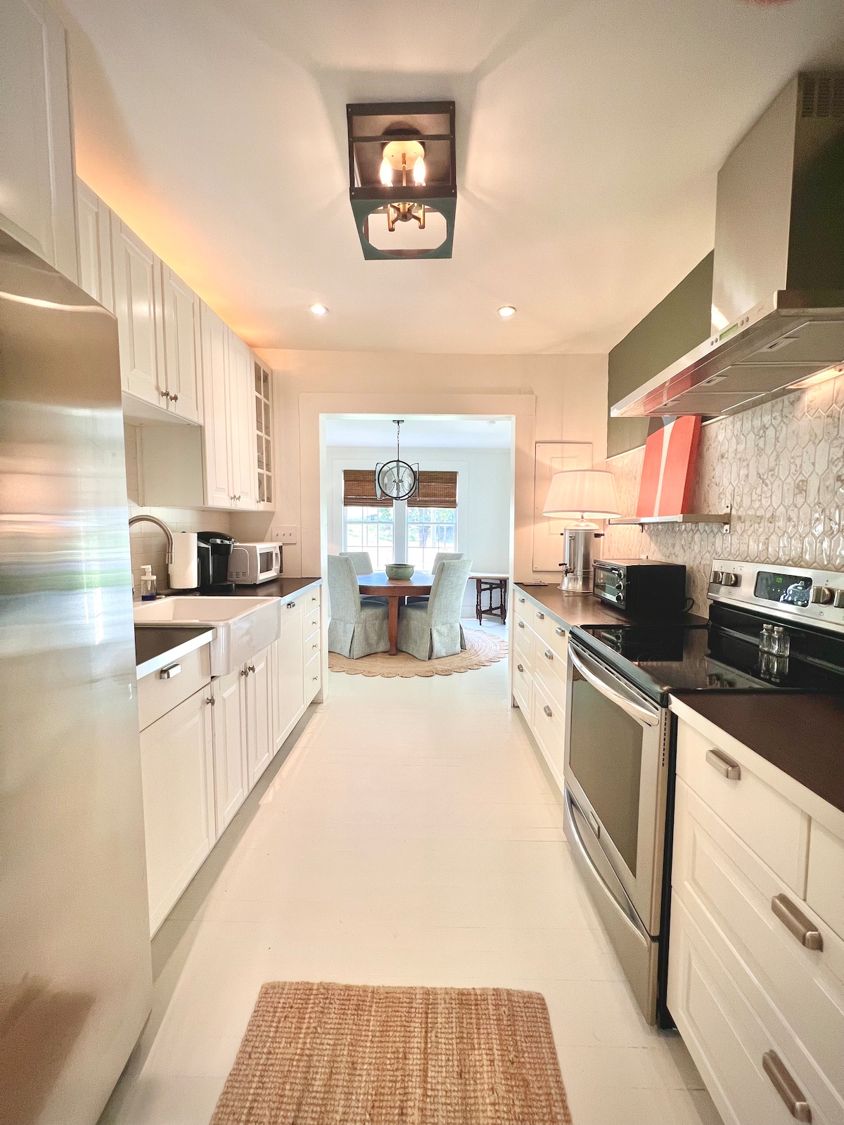 A bright kitchen space features white cabinetry and stainless steel appliances. A narrow runner rug lies on the floor. In the background, a dining area is visible with a round table and four chairs, illuminated by natural light from a nearby window.