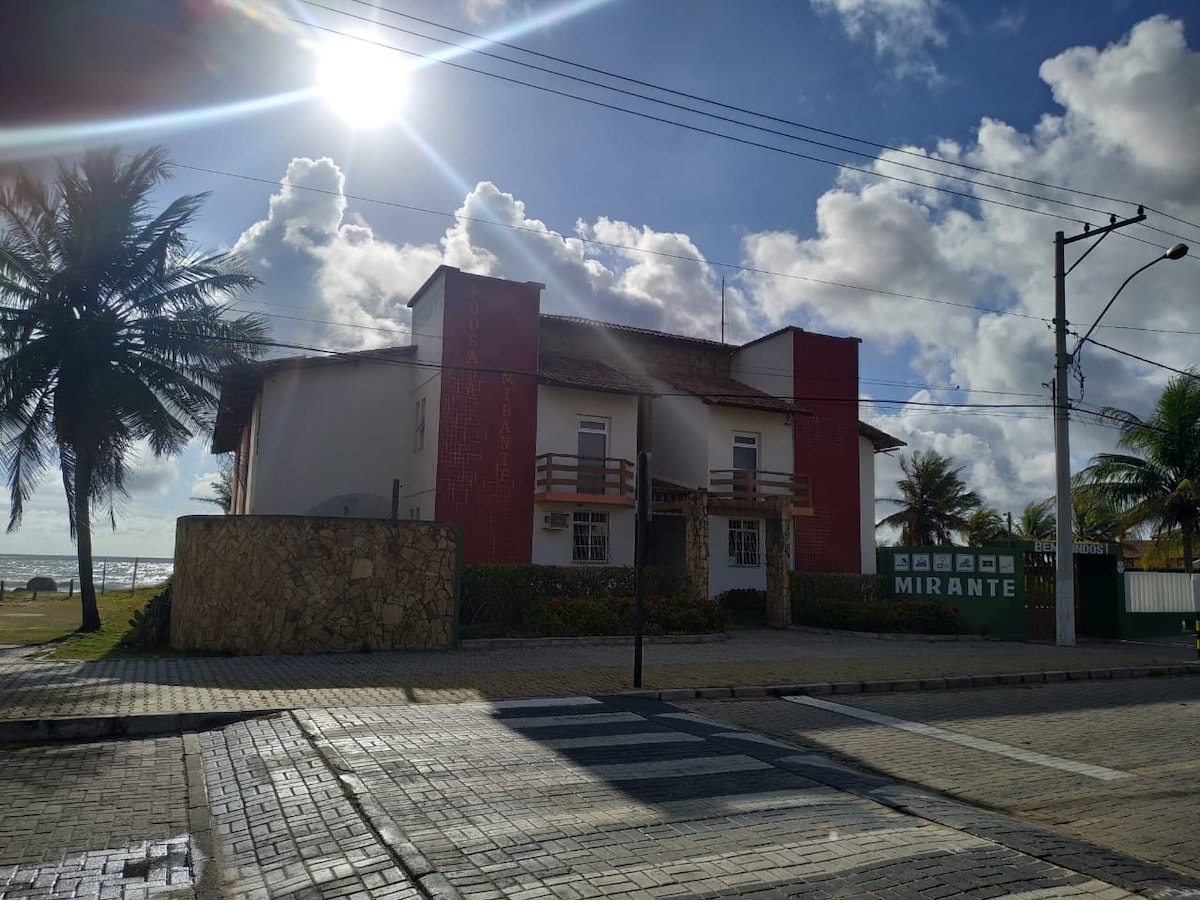 The exterior of a charming lodge is displayed, featuring a two-story structure with a light-colored facade and red accents. Palm trees are visible in the foreground, with a paved pathway leading to the entrance. The bright sun shines above, adding warmth to the scene.