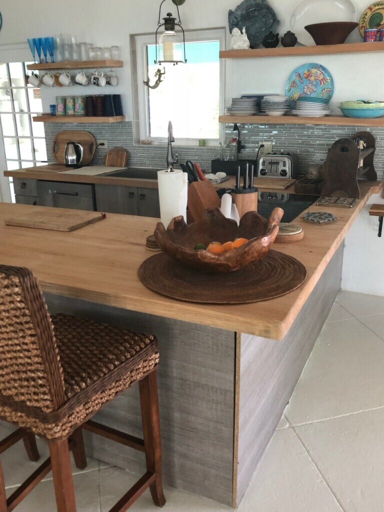 A modern kitchen is showcased with sleek cabinetry and a large wooden countertop. A bowl of colorful fruits sits at the center, while various utensils and tableware are neatly arranged on open shelves and countertops. A wicker chair is positioned at the bar height area.