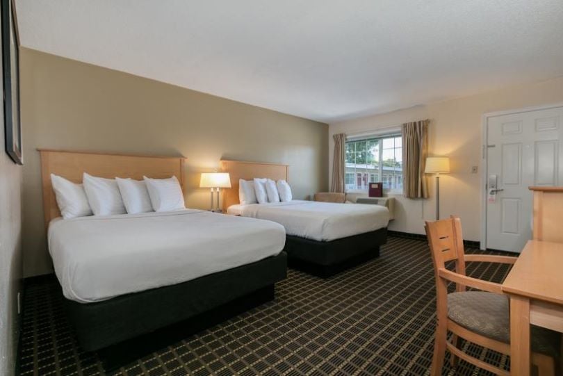 A spacious guest room features two large beds with crisp white linens and plush headboards. A wooden desk and chair are situated beside a window adorned with neutral curtains. A bedside lamp provides soft lighting, while a door leads to the exterior.