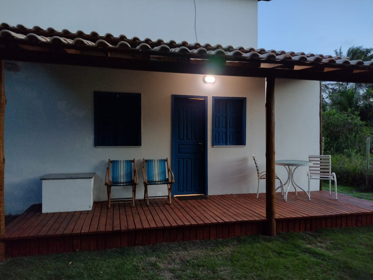 A front porch is shown with wooden decking, featuring two striped chairs alongside a small round table. A blue door is positioned centrally, flanked by two windows. Soft lighting highlights the entrance, creating a welcoming appearance in the evening.