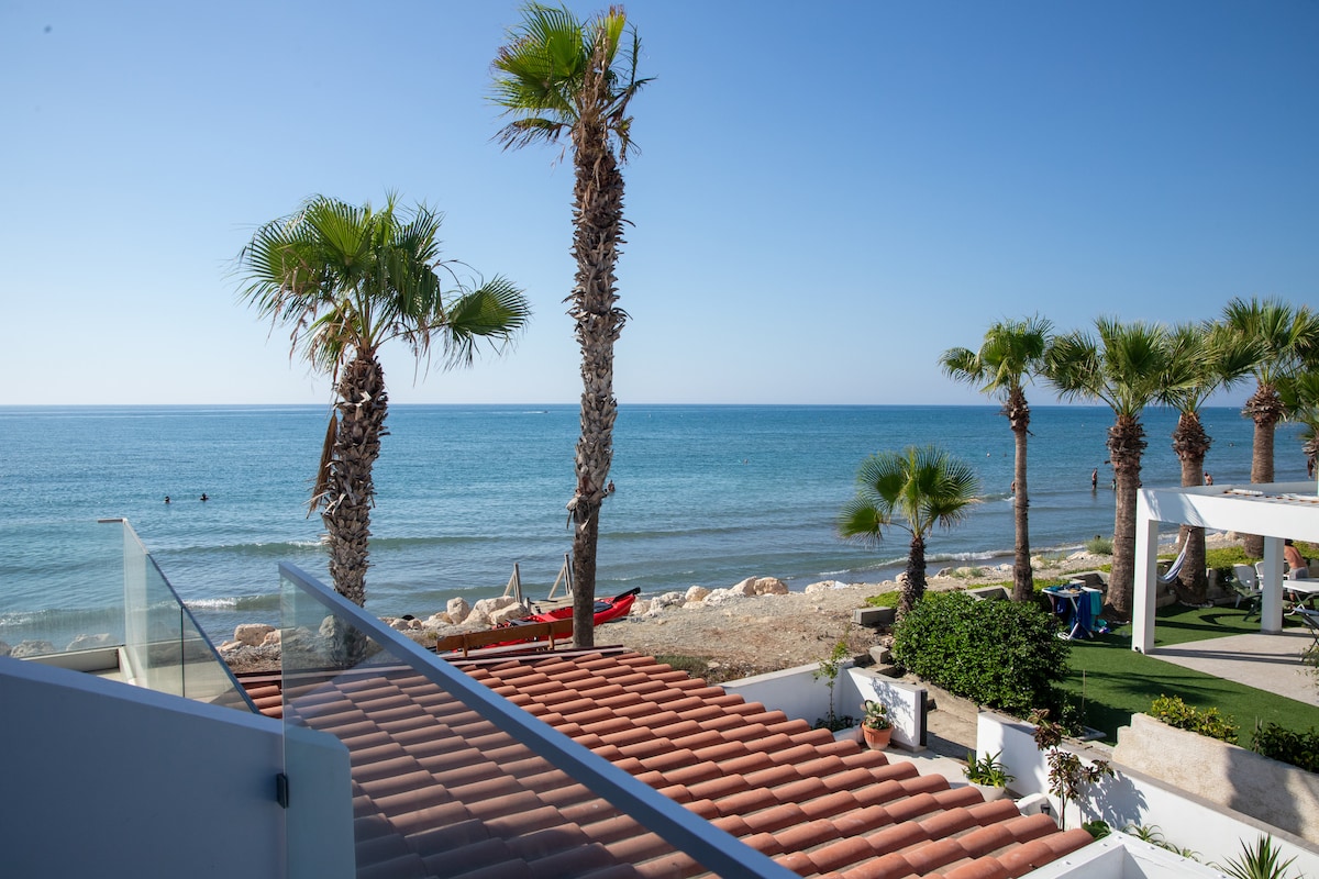 A clear view of the Mediterranean coastline is presented, featuring palm trees that line the beach. Gentle waves lap at the shore, and distant figures enjoy the water. The vibrant blue sky enhances the serene scene, complemented by the terracotta-tiled roof in the foreground.