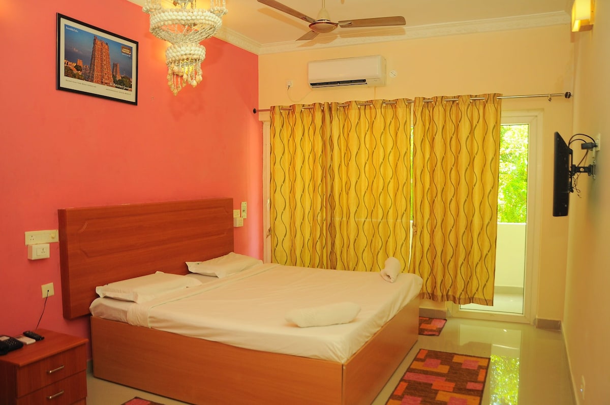 A well-lit bedroom features a spacious bed with crisp linens, complemented by a colorful accent wall and decorative rug. Sheer yellow curtains frame a sliding glass door leading to a small balcony, while a ceiling fan and framed artwork add inviting touches.