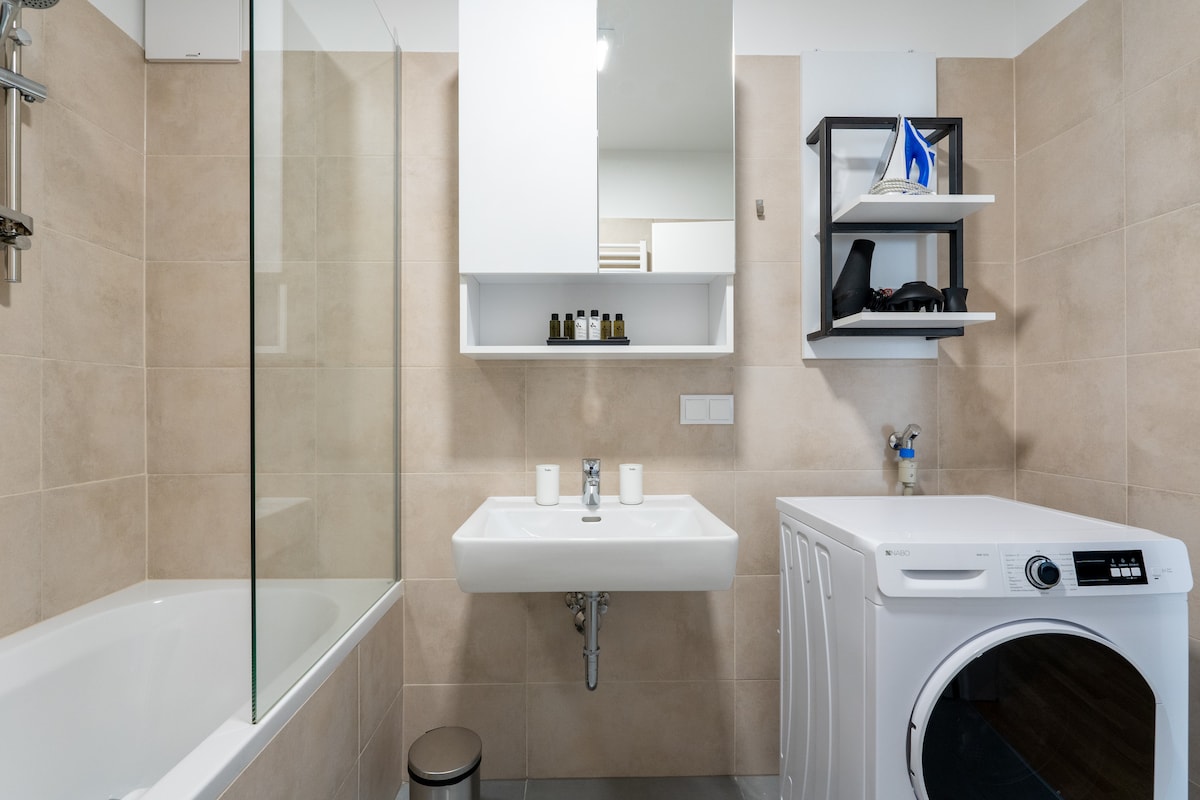 A modern bathroom features a sleek bathtub alongside a contemporary sink. Above the sink, shelves hold toiletries and decorative items. A washing machine is situated beside the sink, with light-colored tiles enhancing the space's clean aesthetic. Natural light is welcomed through a glass enclosure.
