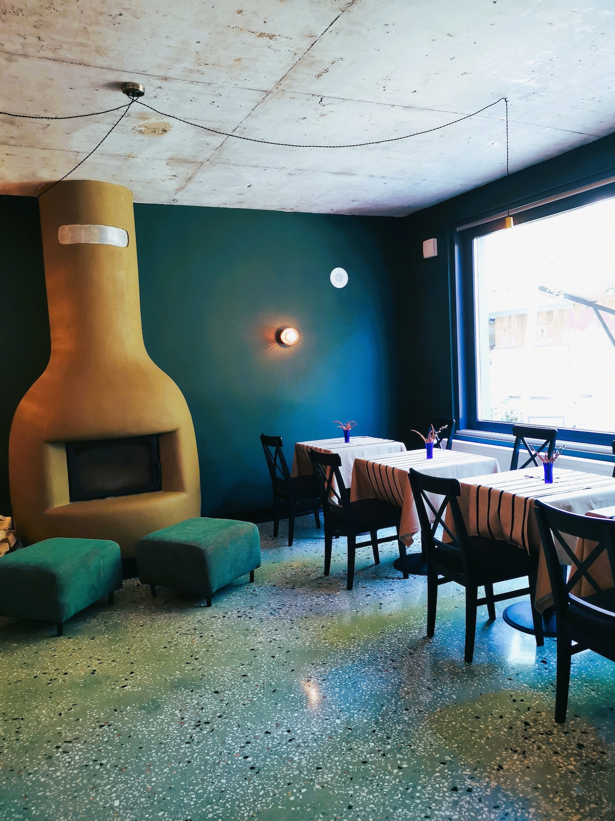 A dining area is illuminated by natural light streaming through a large window. A unique, bottle-shaped fireplace serves as a focal point against a green wall. Tables are set with subtle tablecloths, while several chairs provide seating around them, complemented by two cushioned stools.