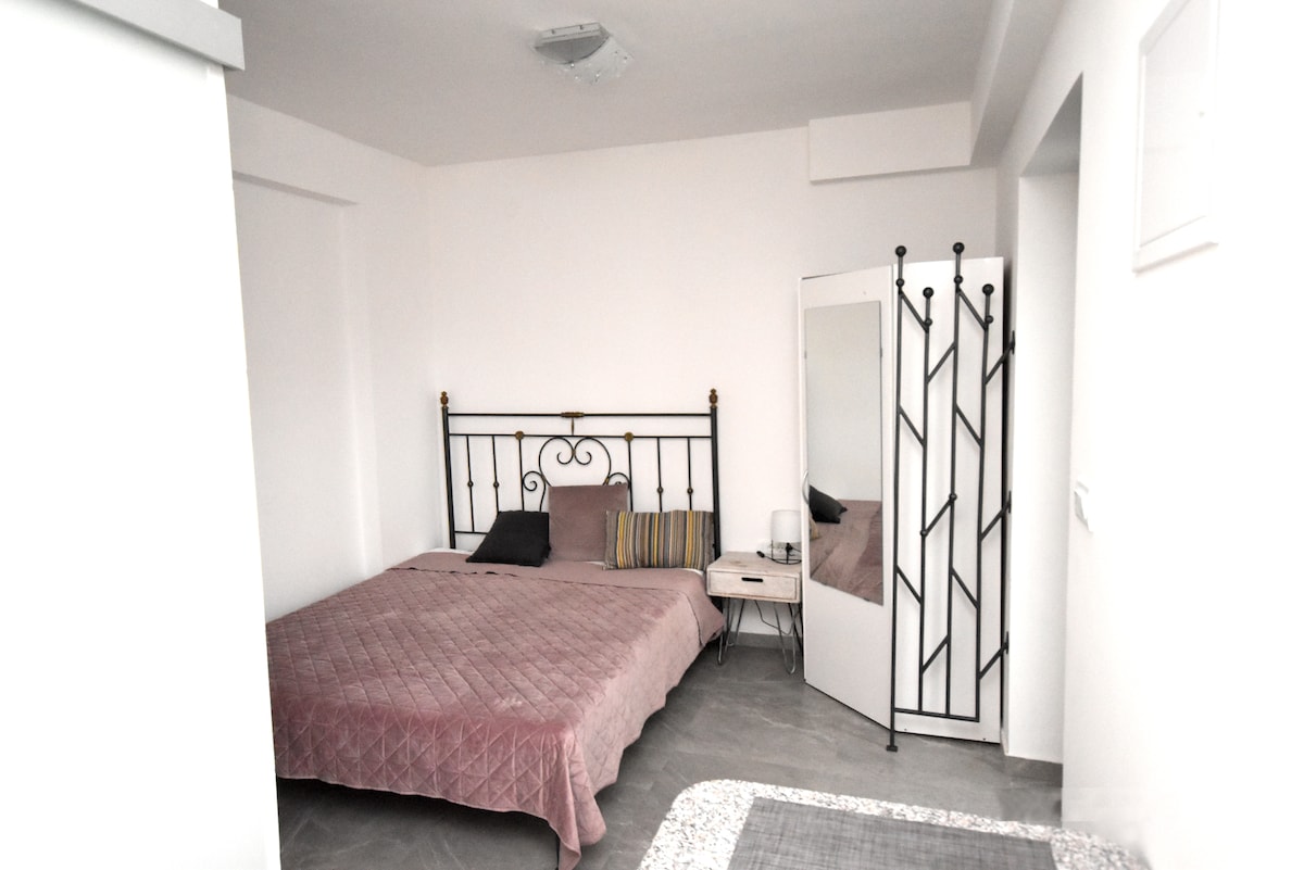 A spacious room features a metal frame bed covered with a soft quilt in muted tones. A nightstand sits next to the bed, accompanied by a decorative mirror and a floor screen. Natural light fills the area, enhancing the simple, modern aesthetic.