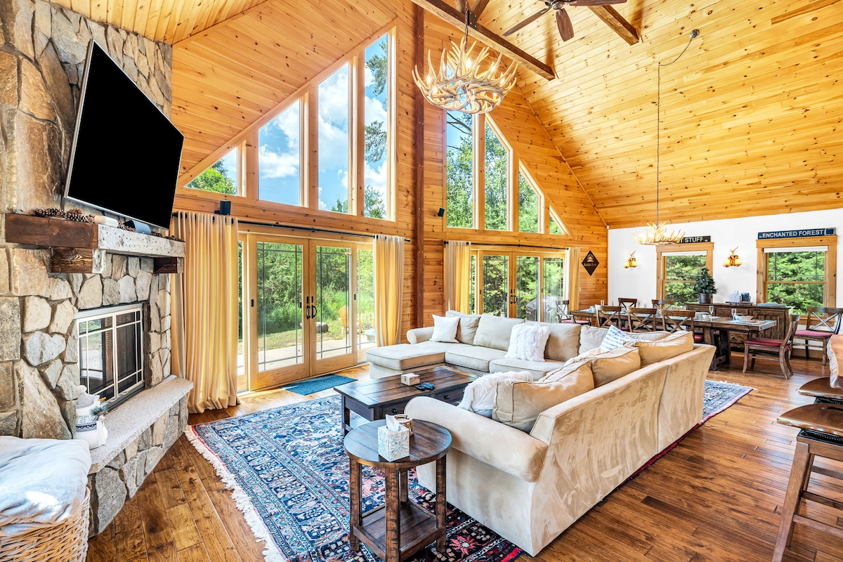 A spacious living area features a large sectional sofa facing a stone fireplace. Floor-to-ceiling windows provide abundant natural light and views of the outdoor greenery. Wooden beams and walls enhance the cozy ambiance, while a patterned area rug adds warmth to the hardwood flooring.