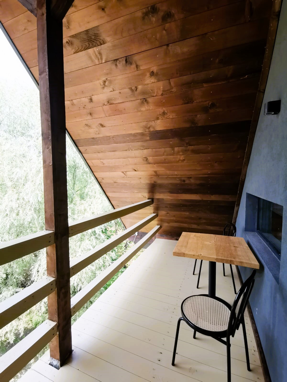 A wooden balcony offers a serene outdoor space, featuring a small table accompanied by two black chairs. The slanted wooden roof adds architectural interest while surrounding greenery provides a natural backdrop.