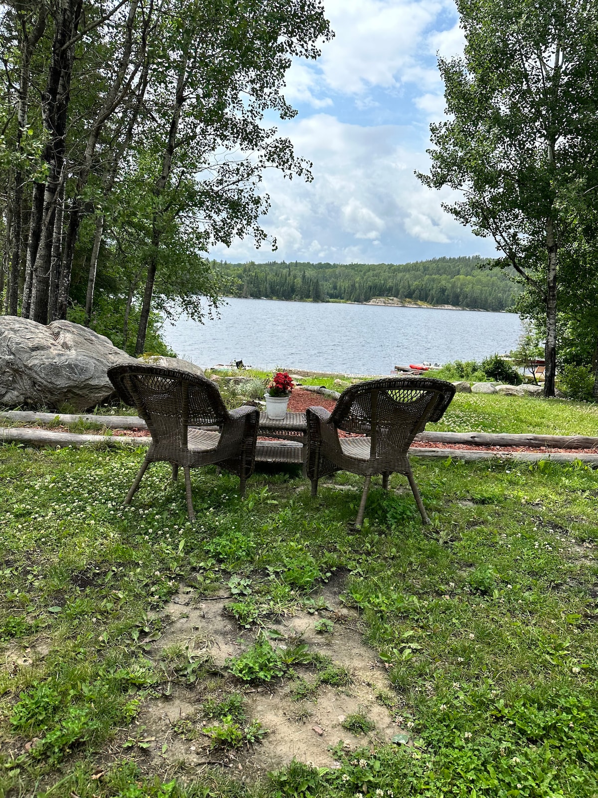 Two wicker chairs are positioned on a grassy area, facing a serene lake surrounded by trees. A small table is placed between them, and a blooming flower arrangement adds a touch of color. Gentle hills are visible in the background.