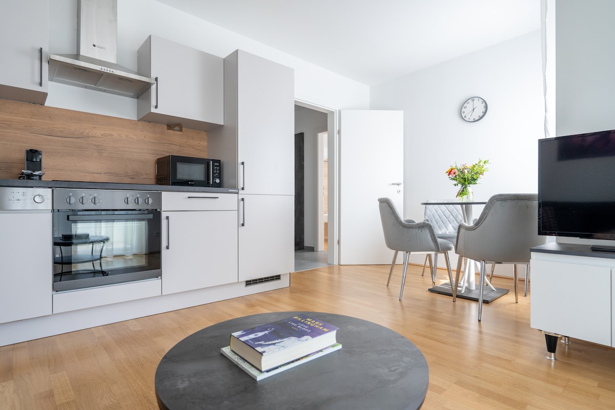 The open kitchen boasts modern appliances including a stove and microwave, paired with light cabinetry. A dining area with two chairs is positioned beside a round coffee table, adding functionality to the space. A wall clock and television are seen in the background.