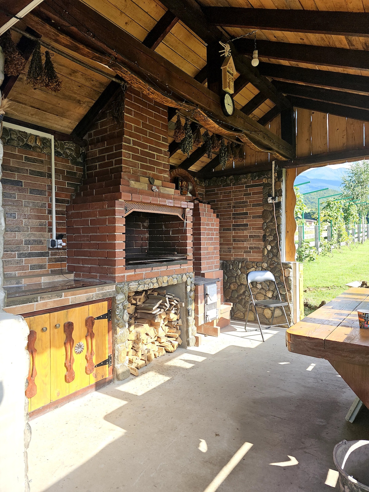 An outdoor grilling area is presented, featuring a sturdy brick barbecue built into a rustic wooden structure. A stacked pile of firewood is neatly arranged nearby. To the side, a spacious dining table surrounded by chairs invites group gatherings, while greenery is visible in the background.