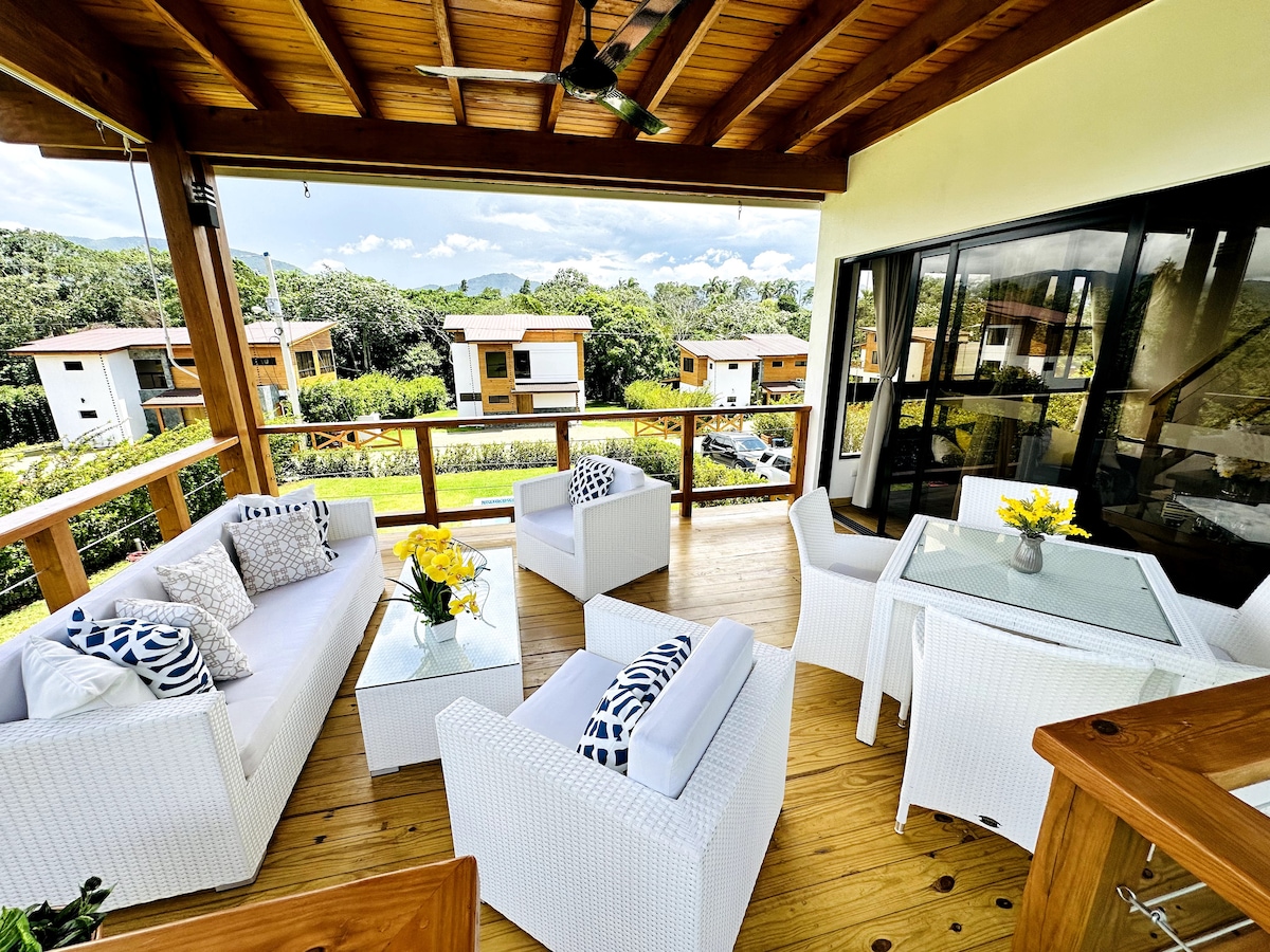 A spacious terrace features comfortable white lounge furniture, including two armchairs and a sofa, arranged around a glass table. Decorative cushions add a touch of style. Lush greenery and mountain views are visible through large glass doors and windows.
