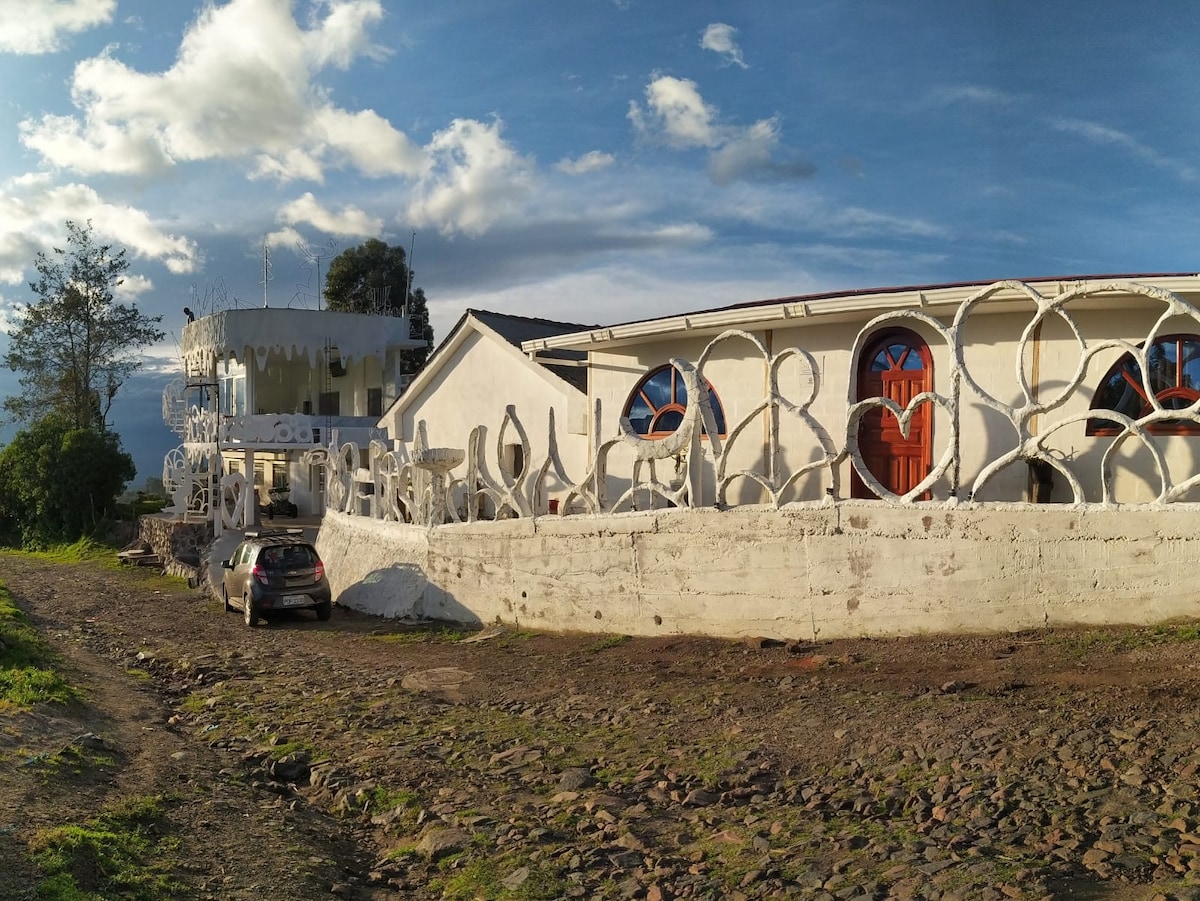 A unique building showcases a blend of traditional and modern architecture, featuring round windows and decorative exterior walls. A gravel path leads to the entrance, with a car parked nearby. Lush greenery and mountains are visible in the background under a clear blue sky.