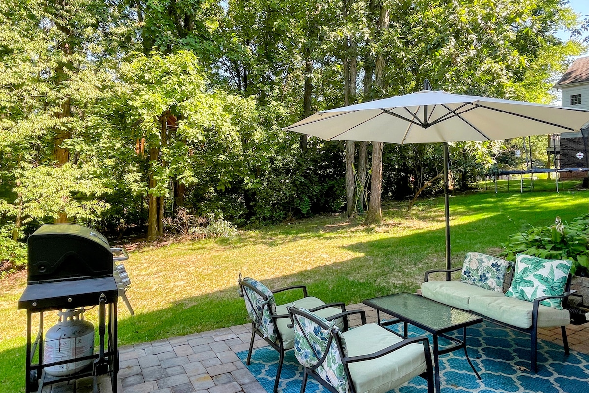 A private patio area is presented, featuring a grill and comfortable seating for relaxing or dining. An umbrella provides shade, enhancing the outdoor experience among lush greenery that offers a serene wooded backdrop.