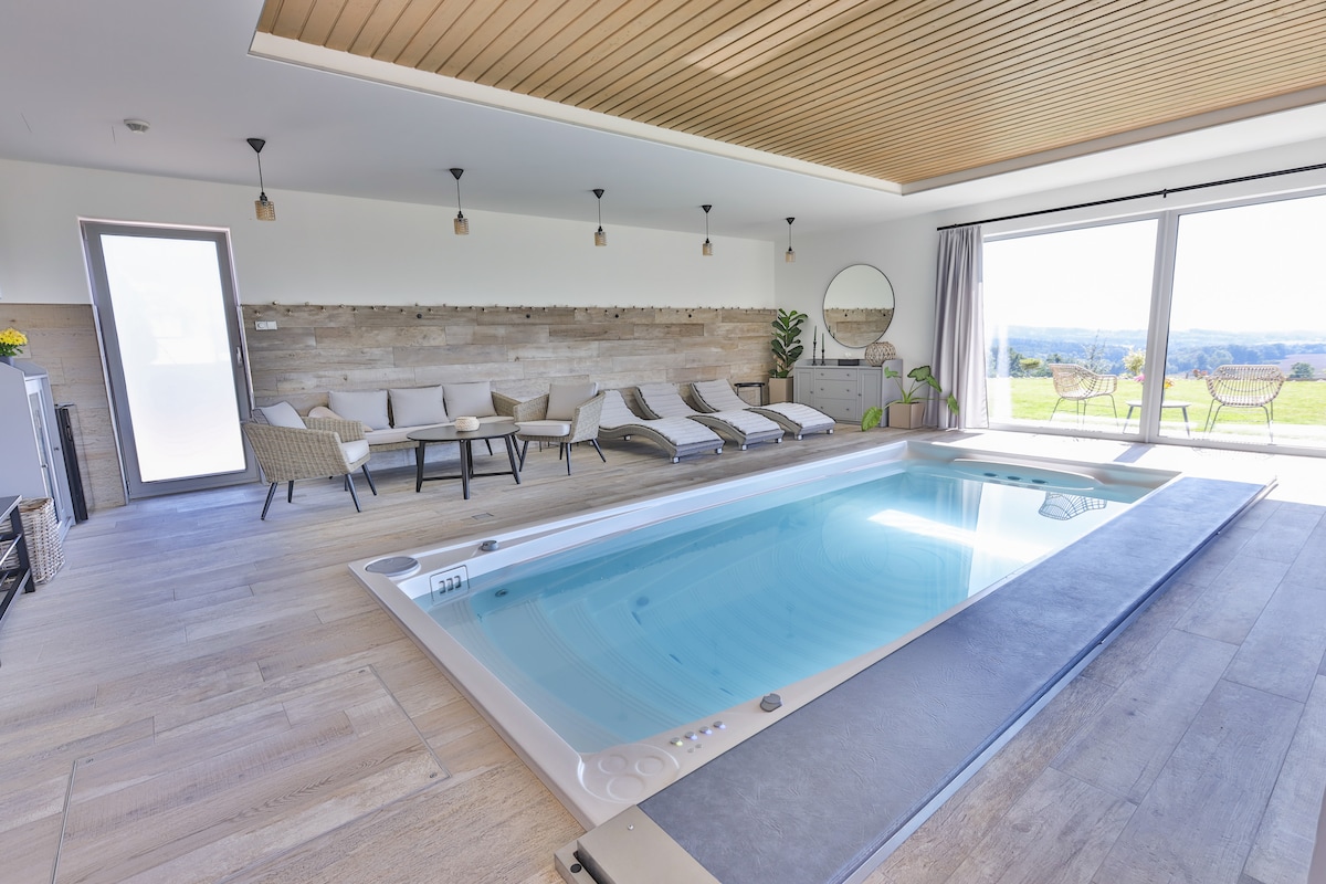 An indoor space featuring a warm, inviting pool surrounded by light wooden paneling. Comfortable loungers are arranged nearby, complemented by large glass doors that offer expansive views of the outdoors. Soft lighting creates a serene atmosphere conducive to relaxation.