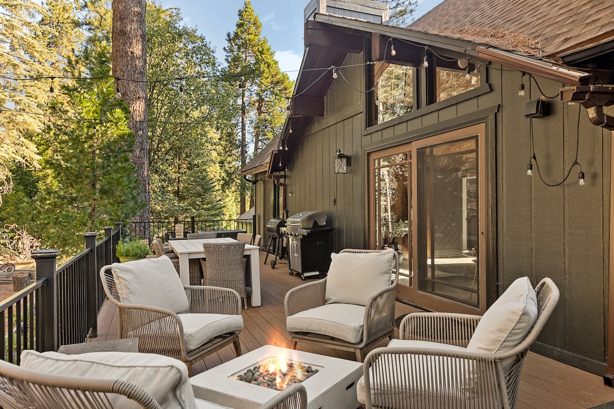 An expansive deck is furnished with a cozy seating area, featuring four cushioned chairs around a fire pit. A BBQ grill is positioned nearby, surrounded by tall trees that enhance the natural setting. Warm string lights provide soft illumination, inviting relaxation outdoors.