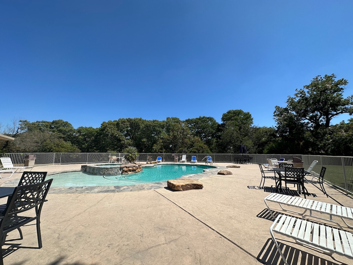 A spacious outdoor pool area is surrounded by a concrete deck. Lounge chairs are positioned around the pool, while a stone feature enhances the space. Lush greenery and trees are visible in the background under a clear blue sky.