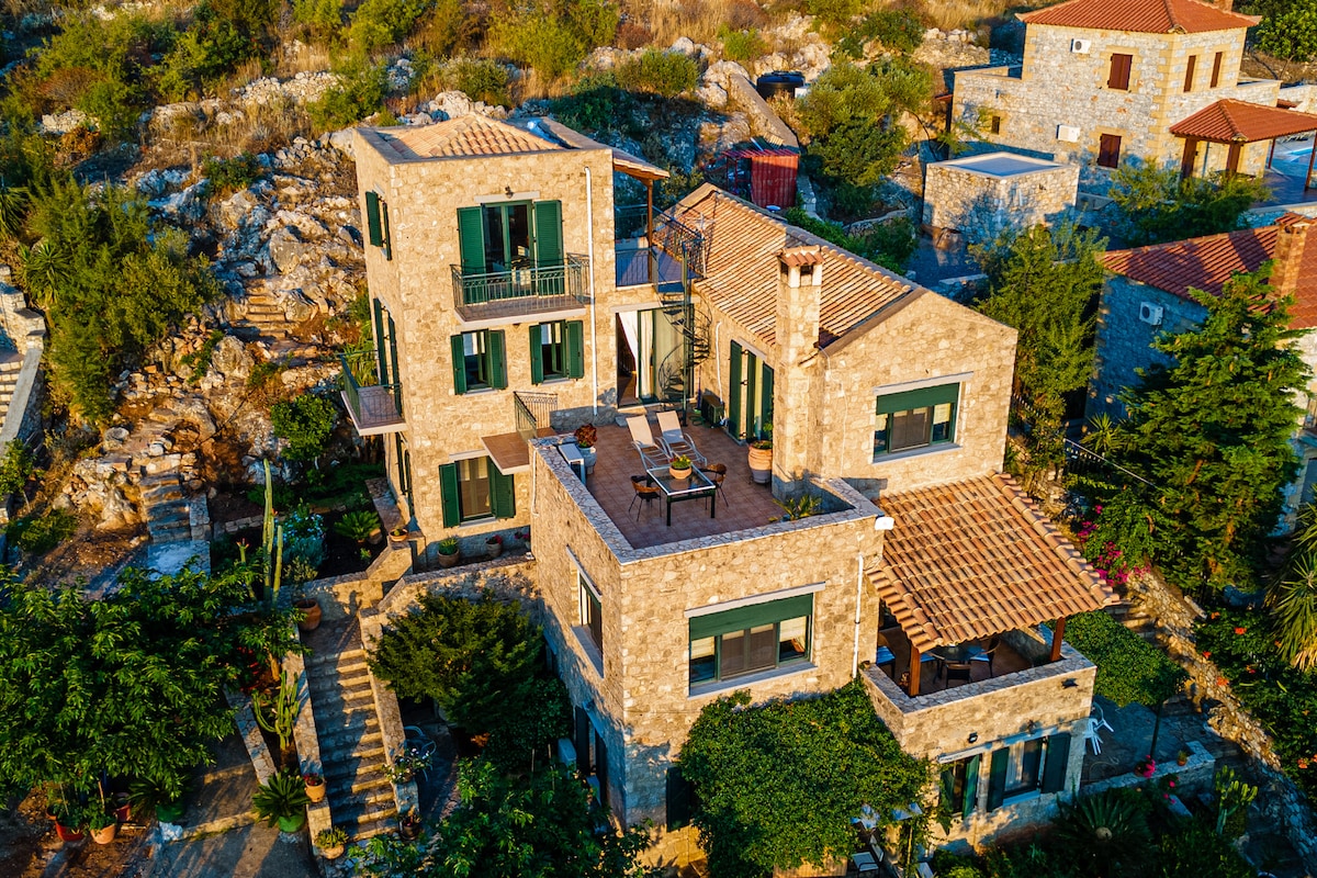 An aerial view of a stone villa showcases multiple levels with green shutters adorning the windows. A spacious terrace features outdoor seating, while a variety of plants enhance the surrounding landscape, providing a warm and natural setting.
