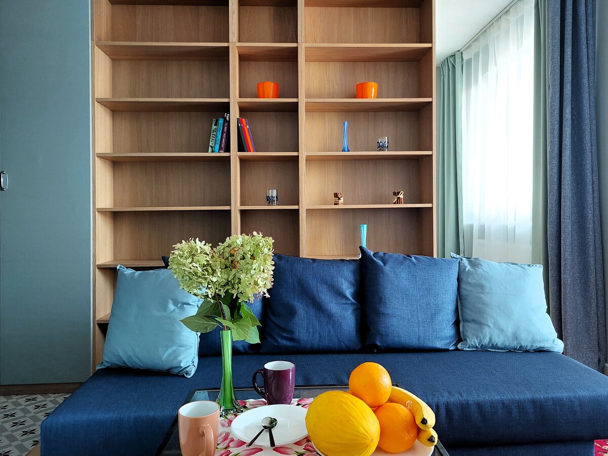 A bright living area showcases a spacious blue sofa adorned with cushions. A coffee table features a floral design, displaying cups and fresh fruits. Behind the sofa, a wooden shelving unit holds various decorative items, enhancing the room's inviting ambiance.