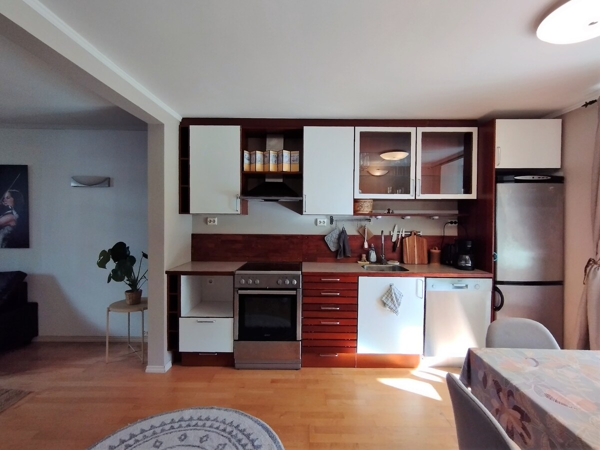 The kitchen area features modern wood cabinetry, a stainless steel oven, and a refrigerator. Countertops display various kitchen utensils and a coffee maker. Natural light is welcomed through the space, enhancing the neutral color palette and creating an inviting environment for meal preparation.