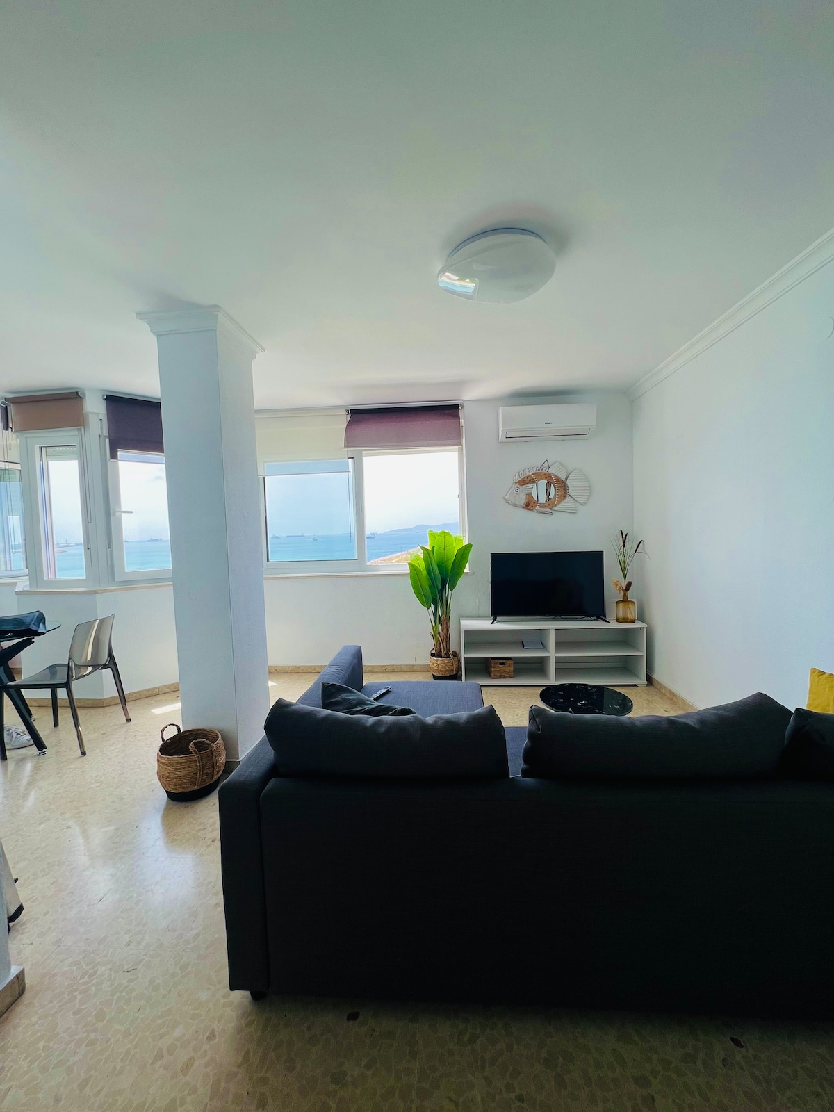 A bright living area features a cozy gray sofa facing a flat-screen television. Large windows allow natural light to fill the space, with views of the outside landscape. A potted plant adds a touch of greenery, and a small basket is placed nearby.