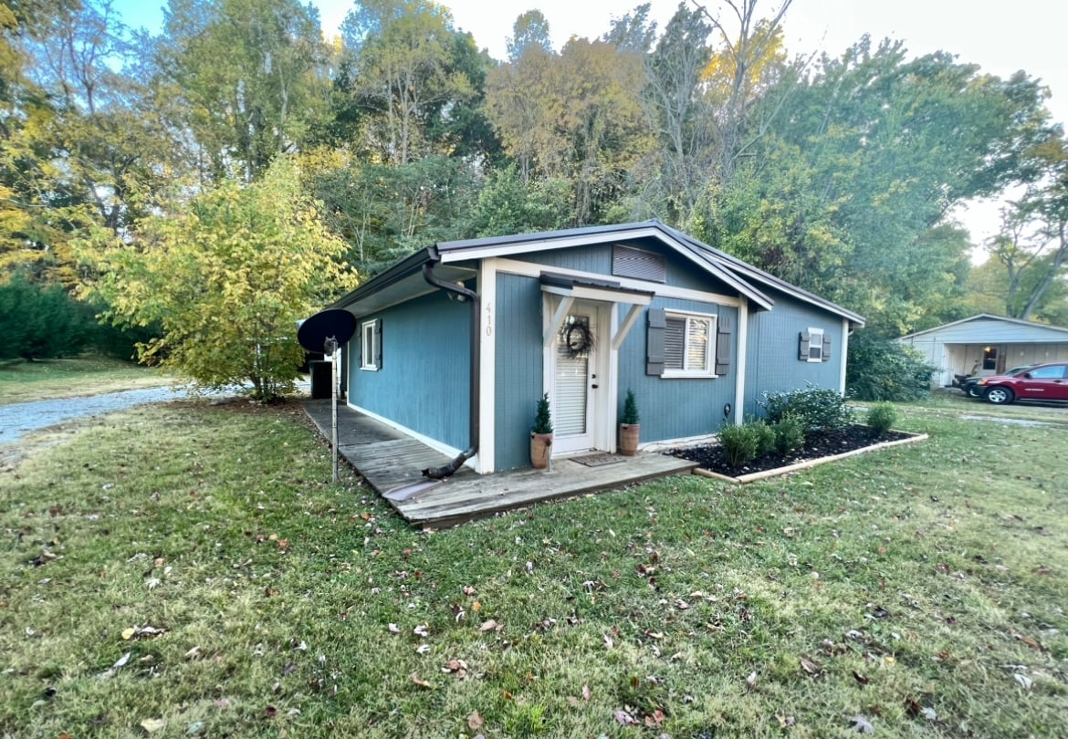 A quaint blue house is framed by greenery and trees. A welcoming front porch is adorned with potted plants, and a wreath is displayed on the door. A gravel pathway leads to the entrance, contributing to the home's approachable character.