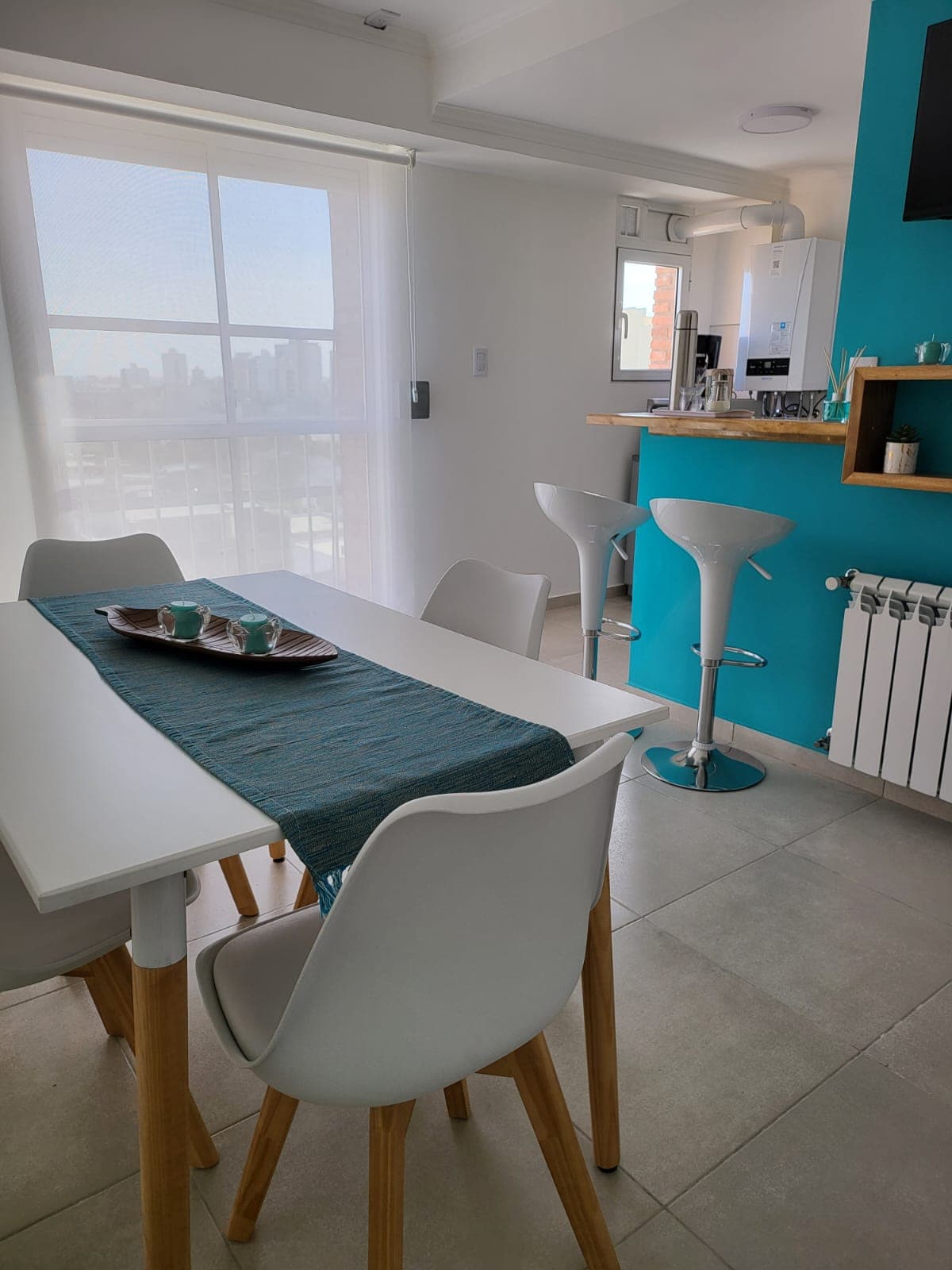 A dining area is highlighted by a white table and four white chairs with wooden legs. A turquoise runner decorates the table, accentuated by a decorative tray. Bar stools are positioned near the kitchen, with a window allowing natural light and city views.